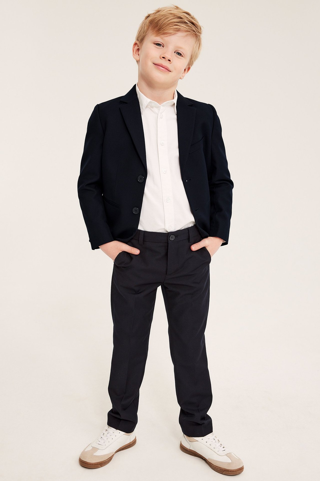 Z przodu, dziecko w granatowych spodniach garniturowych, marynarce, białej koszuli i białych trampkach.