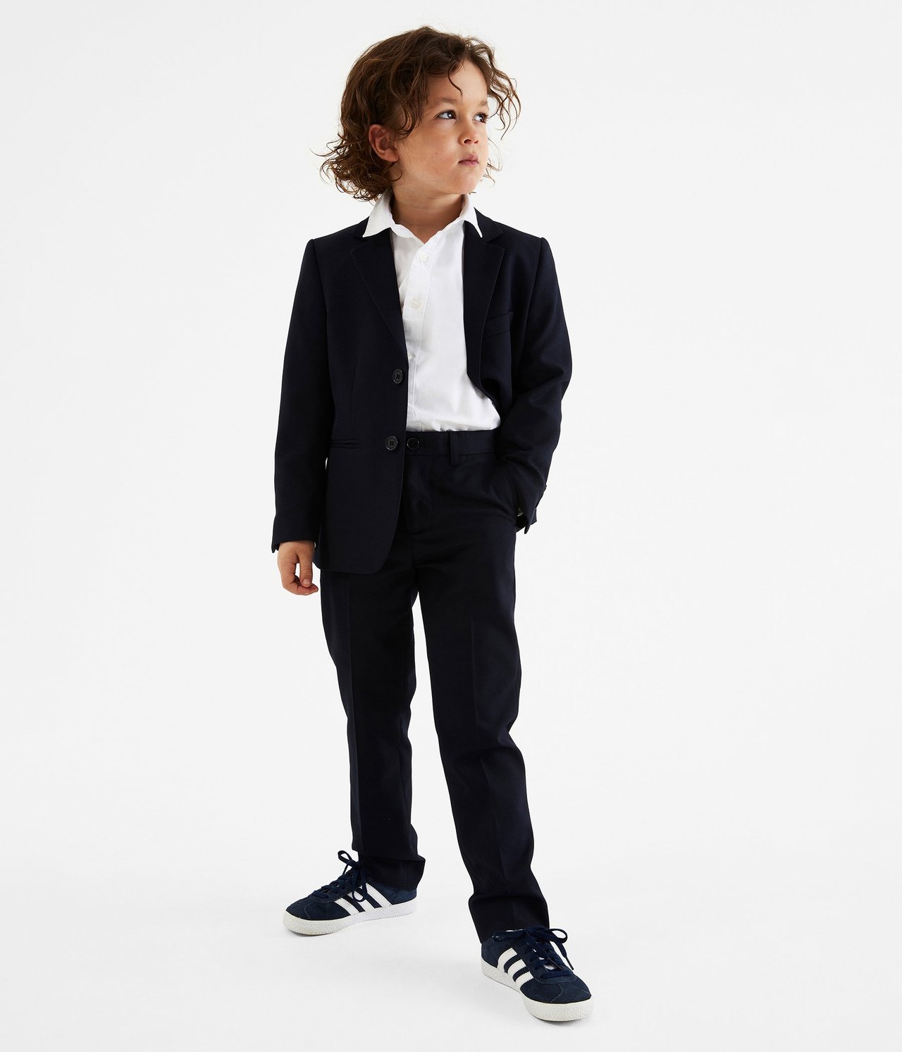 Edestä kuvattu lapsi tummansinisissä puvunhousuissa, bleiserissä, valkoisessa paidassa ja tummansinisissä lenkkareissa.