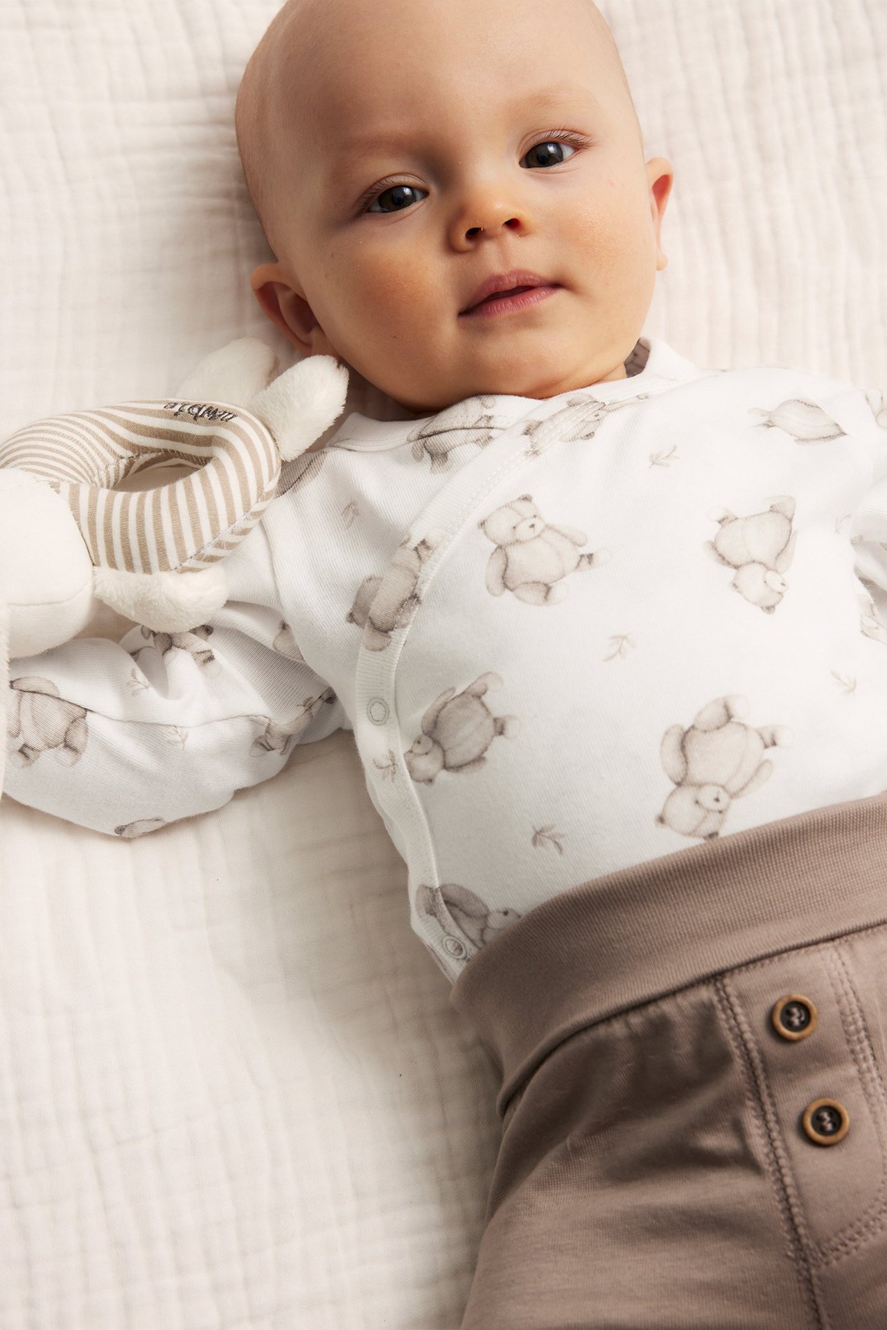 Närbild på baby i offwhite omlottbody med djurmönster, bruna byxor och håller en randig skallra.
