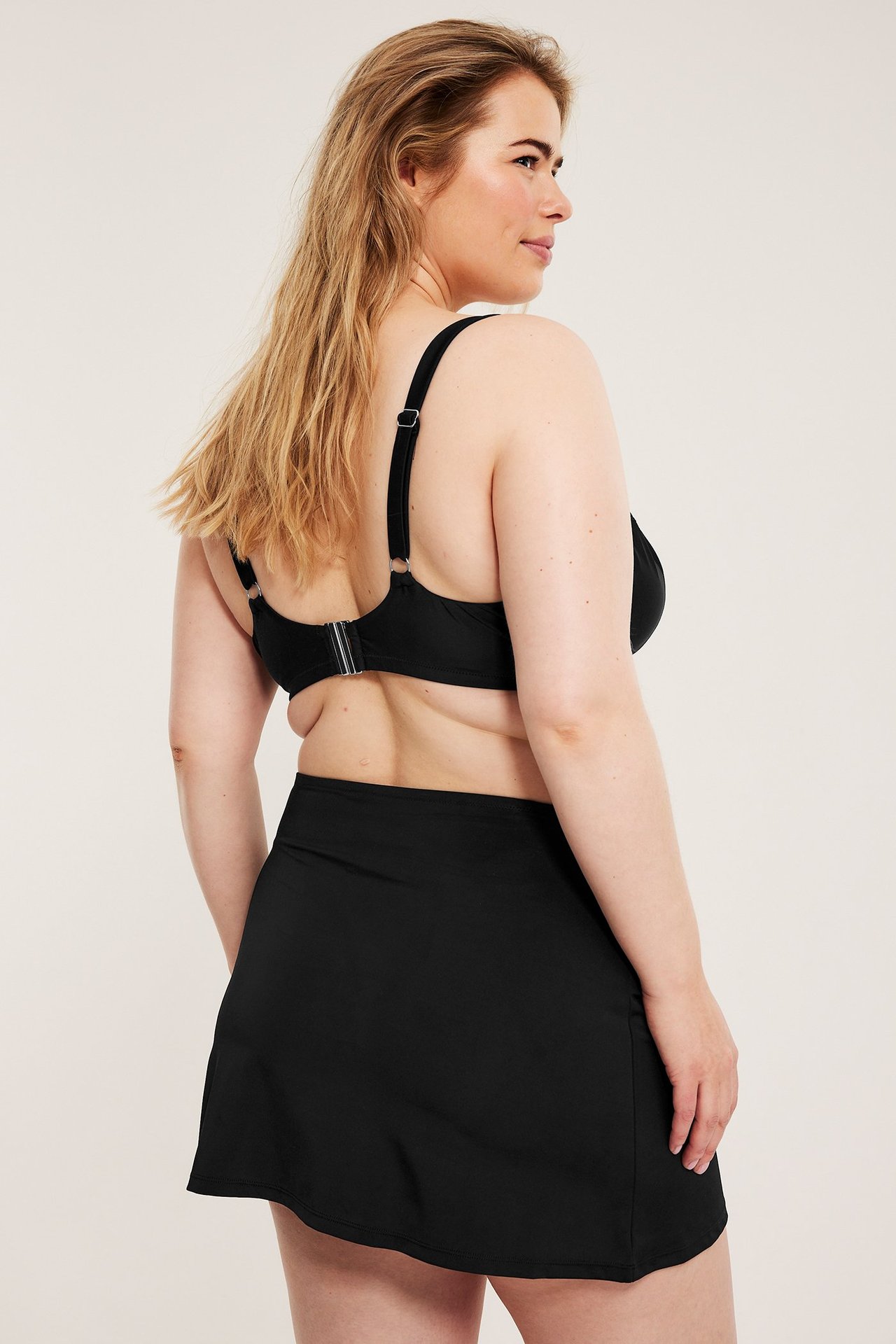 Bakifrån-sidovy av en kvinna i en svart enfärgad utsvängd badkjol och svart bikinitopp.