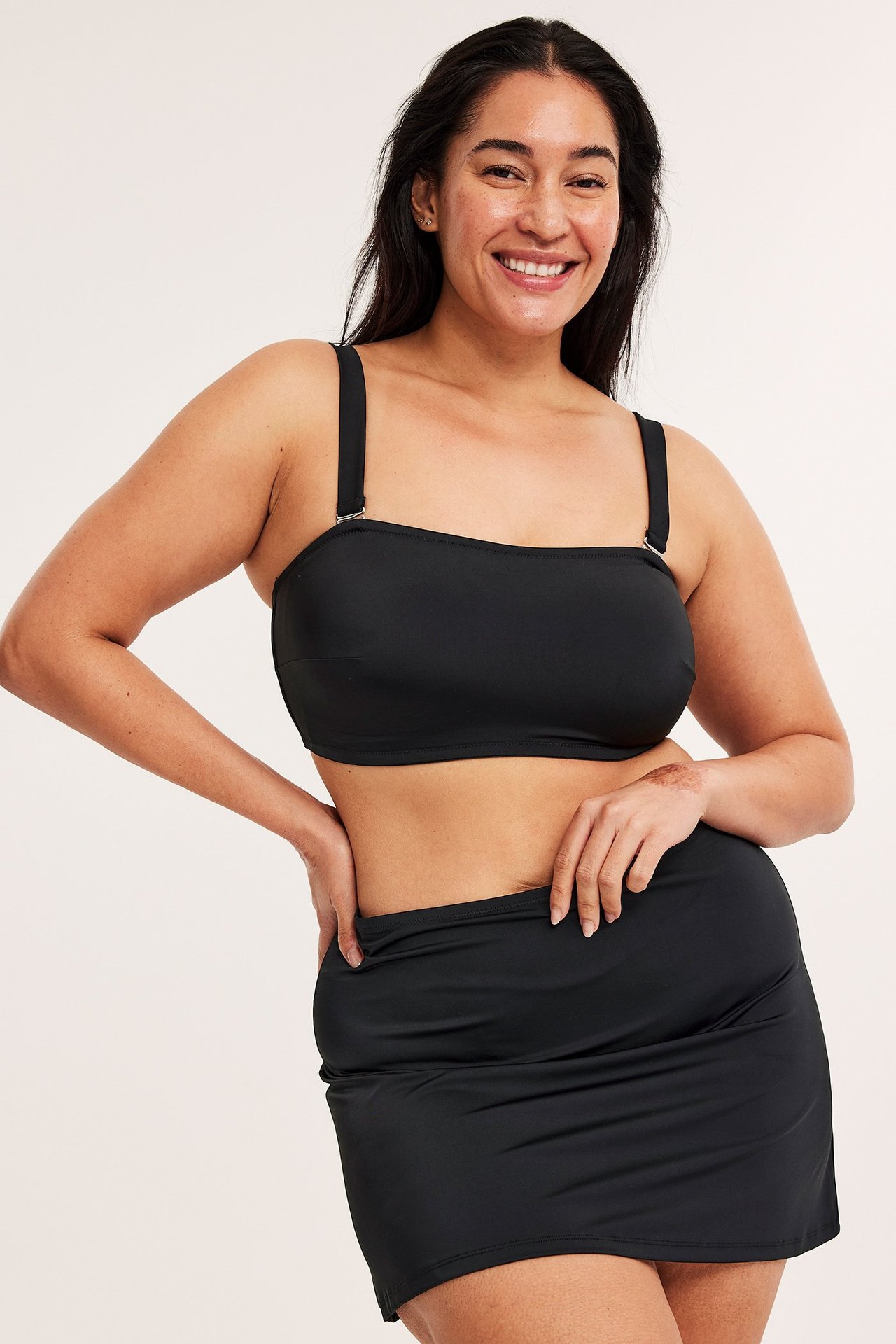 Widok z przodu uśmiechniętej kobiety w czarnej, jednolitej spódniczce kąpielowej i czarnym topie bandeau.
