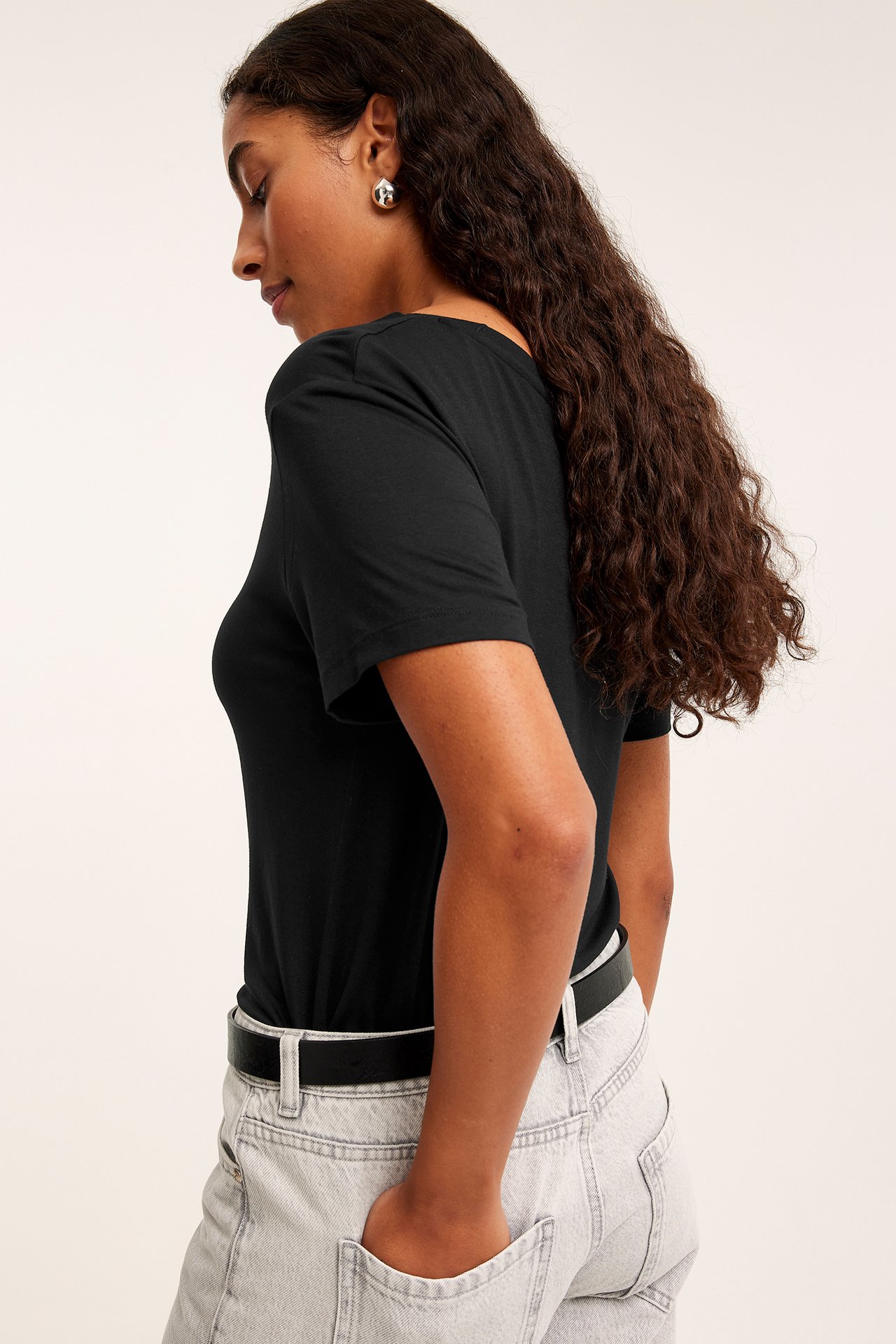 Sidovy av en kvinna i en svart viskos t-shirt med rund hals, ljusgrå jeans, ett svart bälte och silverörhängen.
