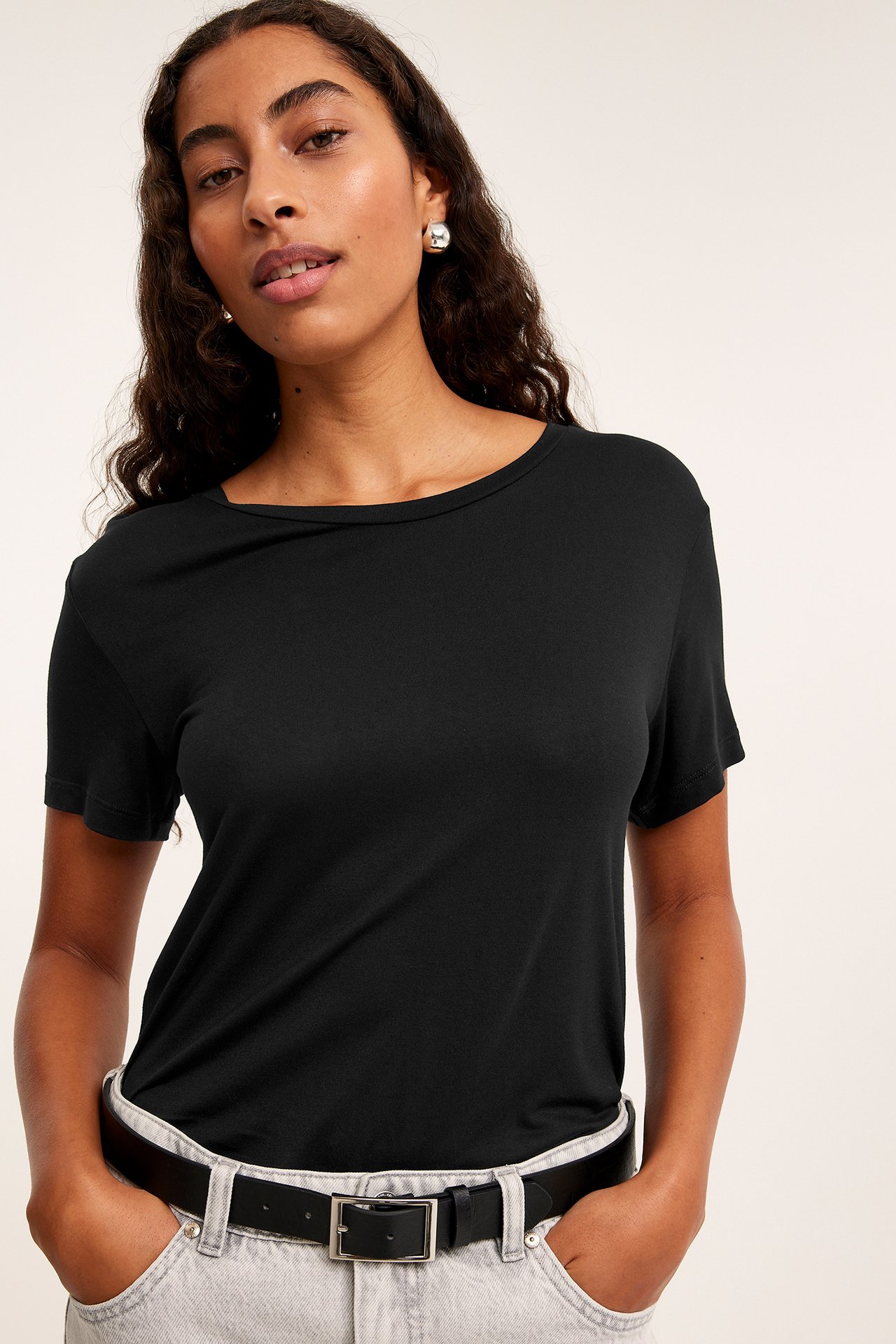 Framsida: Kvinna i svart viskos t-shirt med rund hals, ljusgrå jeans, svart bälte, silverörhängen.