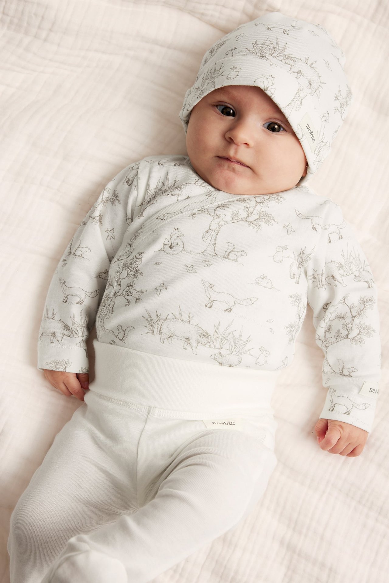 Nærbilde av baby i offwhite mønstret omslagsbody, ensfargede bukser med fot og matchende lue.