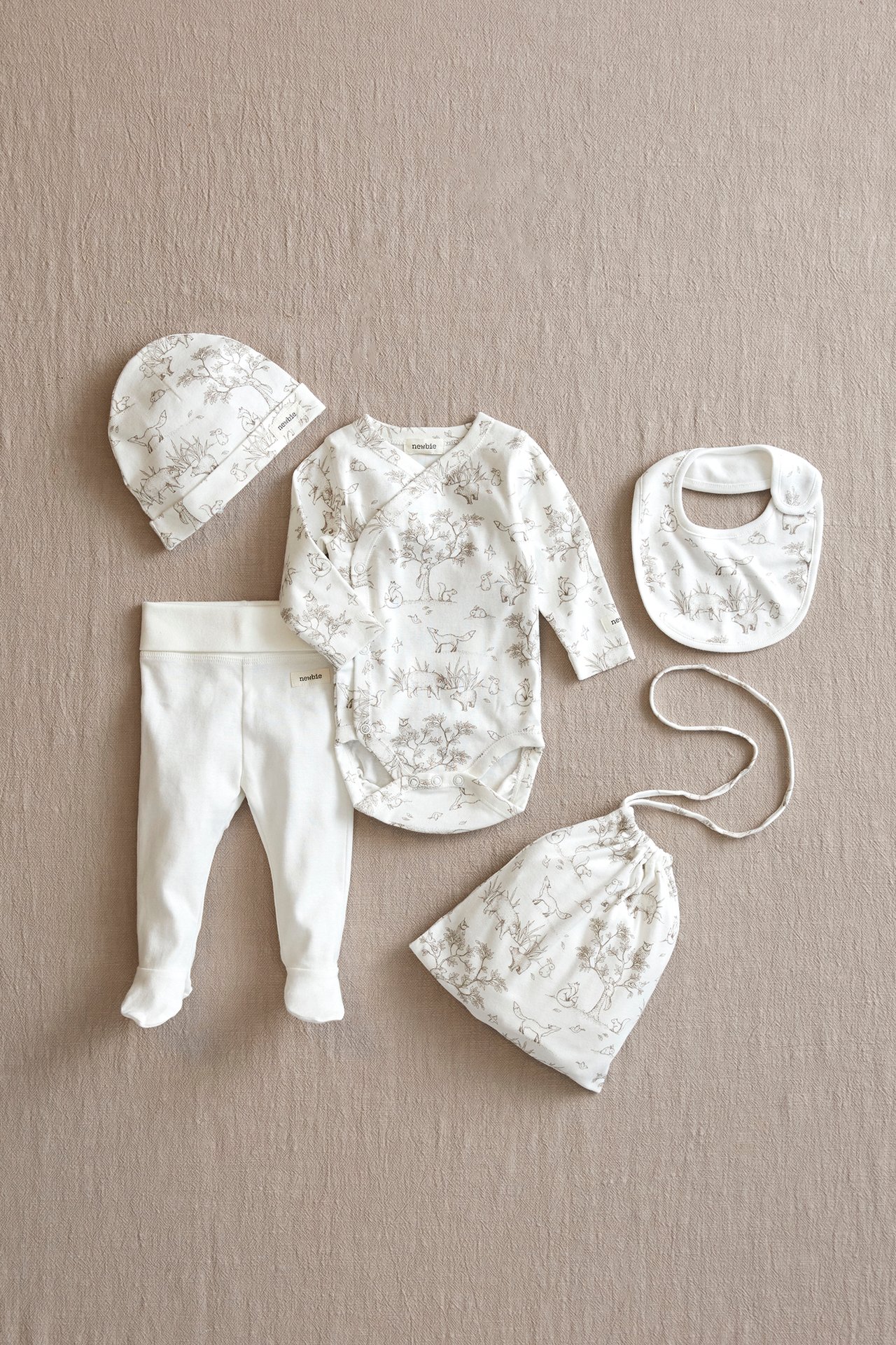 Widok z góry: Ecru wzorzyste body kopertowe dla niemowląt, pasująca czapka, śliniak, worek i jednolite spodnie ze stopkami.