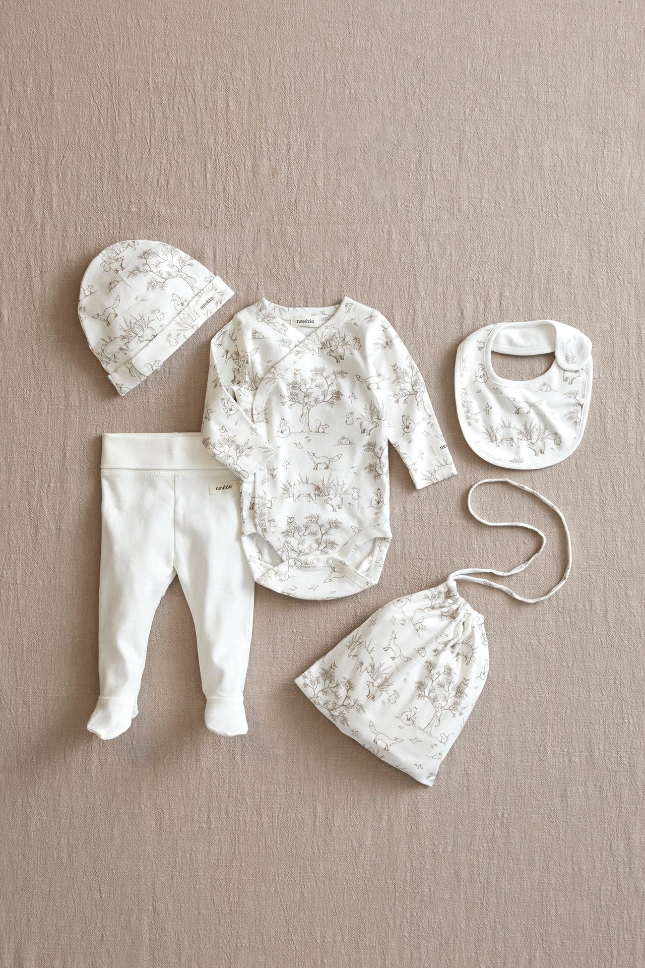 Ovanifrån: offwhite mönstrad babyomlottbody, matchande mössa, haklapp, tygpåse och enfärgade byxor med fot.