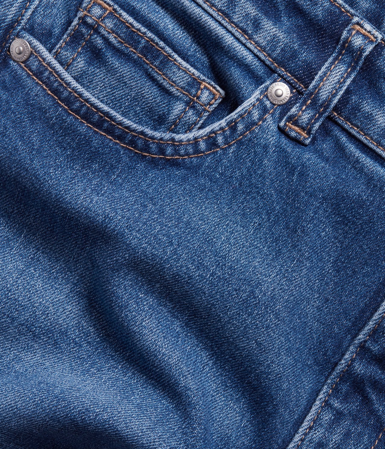 Närbild av blå denim cropped flare jeans med bruna sömmar och silvernitar vid fickan.