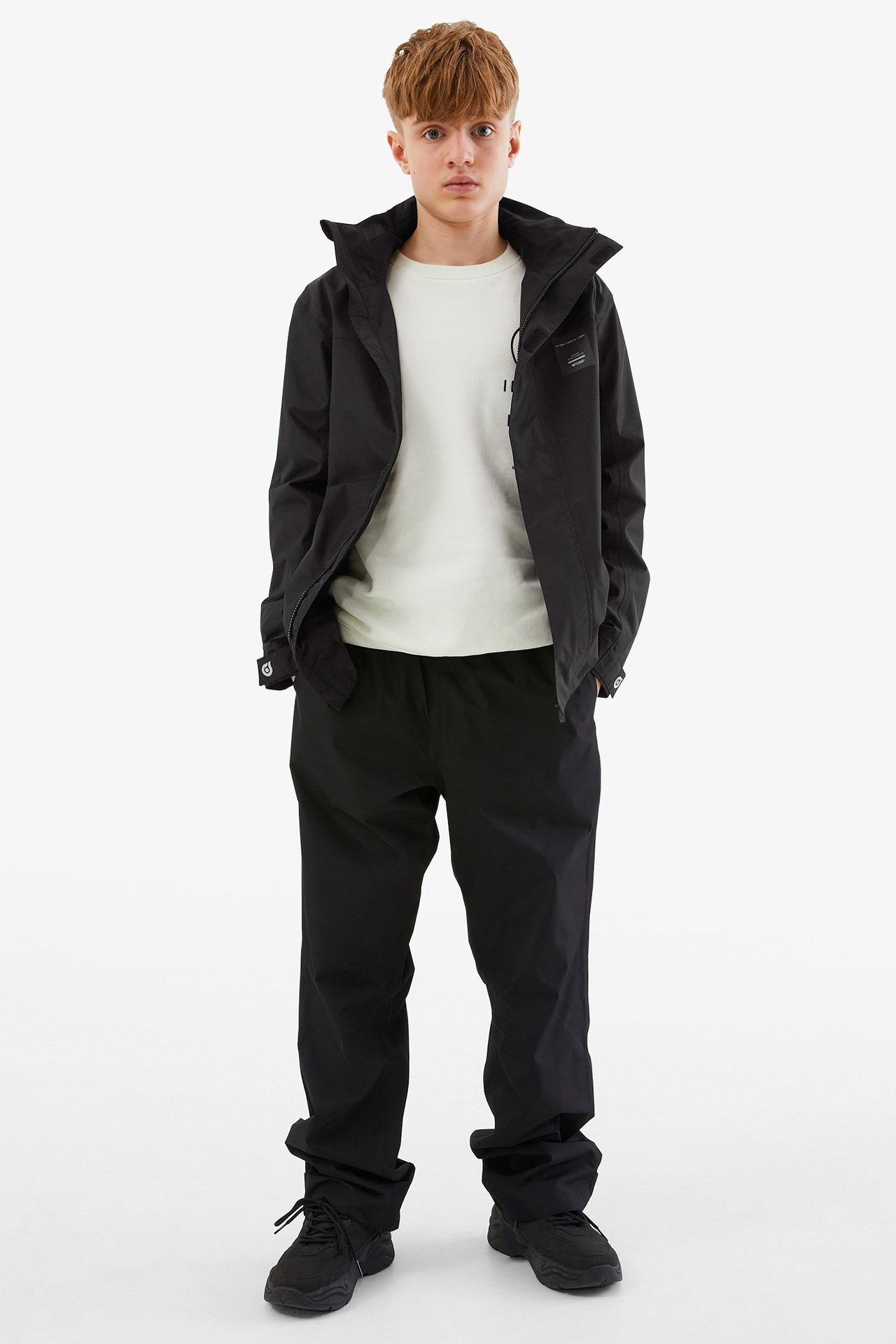 Widok z przodu dziecka w czarnej wodoodpornej kurtce softshell, kremowym T-shircie, czarnych spodniach i czarnych sneakersach.