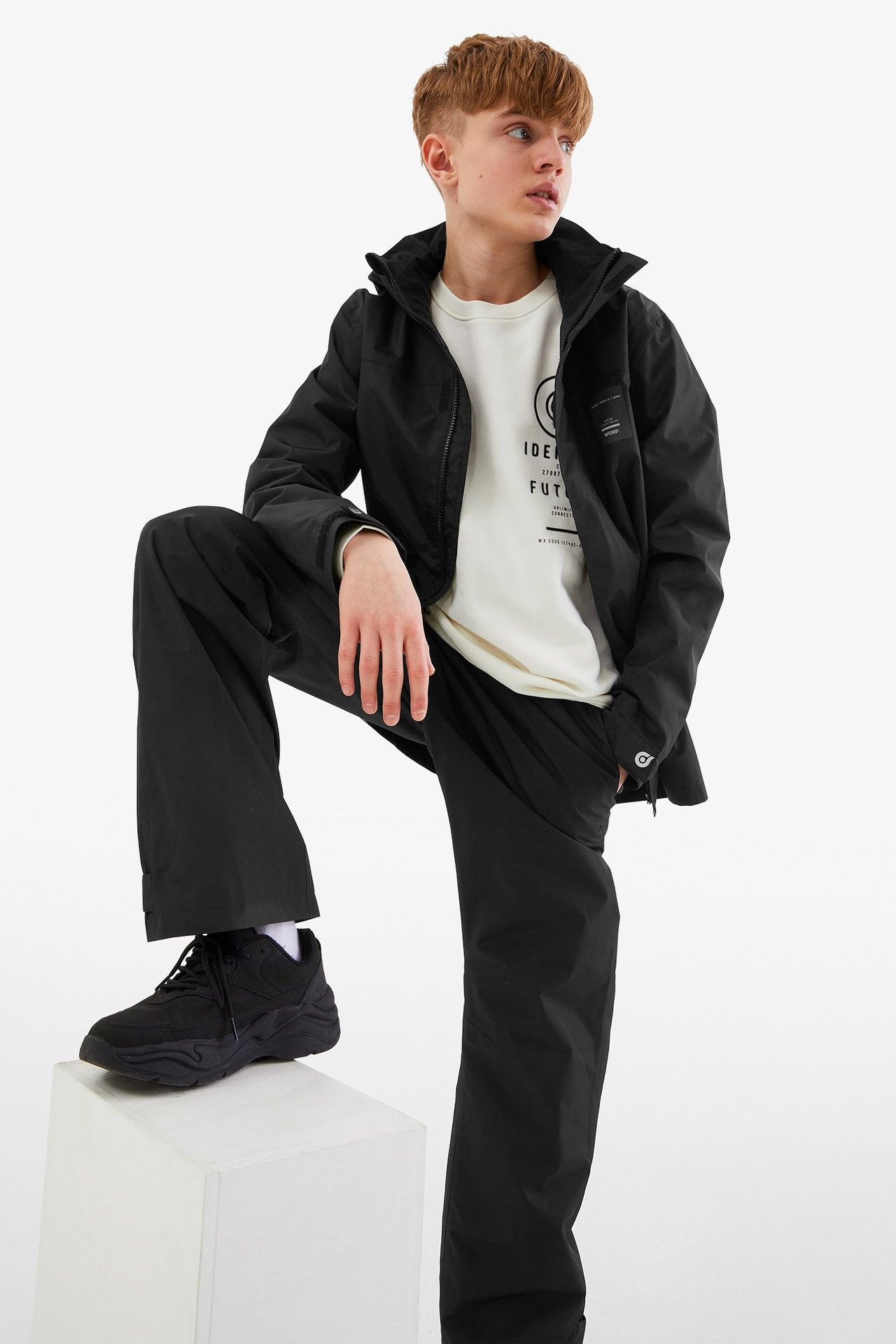 Etunäkymä lapsesta mustissa vedenpitävissä housuissa, mustassa takissa, luonnonvalkoisessa paidassa ja mustissa lenkkareissa.