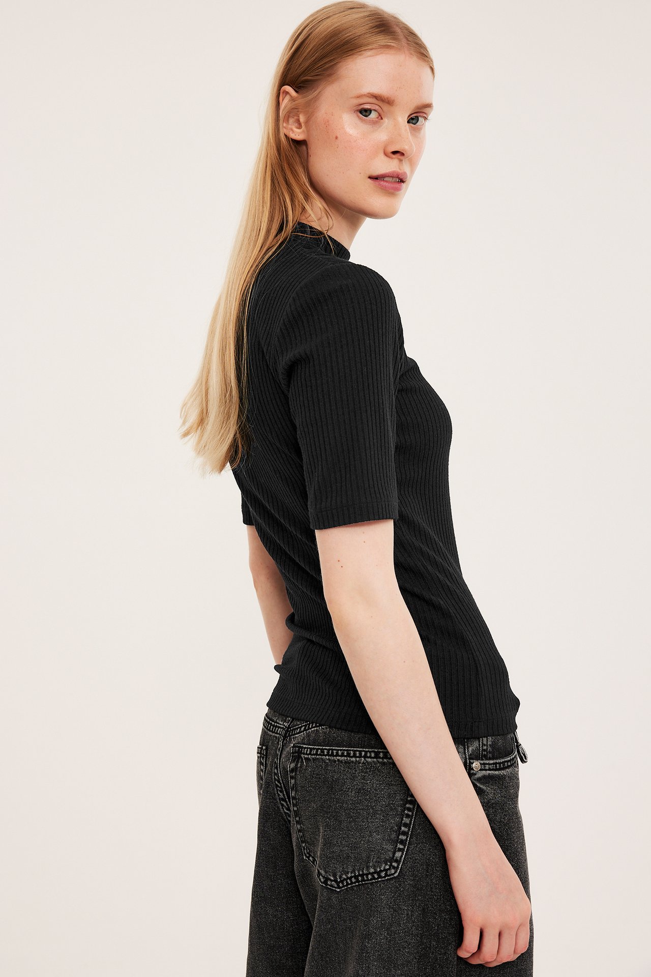 Sidovy av kvinna i svart ribbad kortärmad polotopp och mörkgrå jeans.