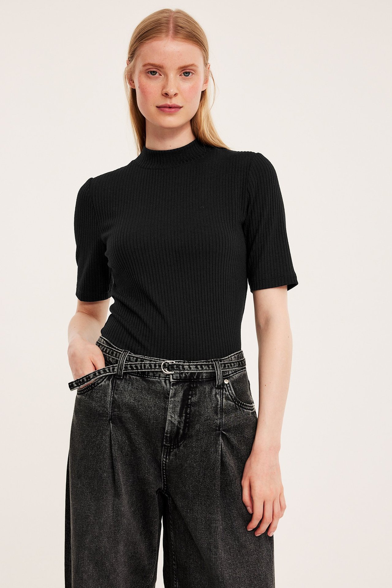Frontvy av kvinna i svart ribbad kortärmad polotopp och mörkgrå vida jeans med bälte.