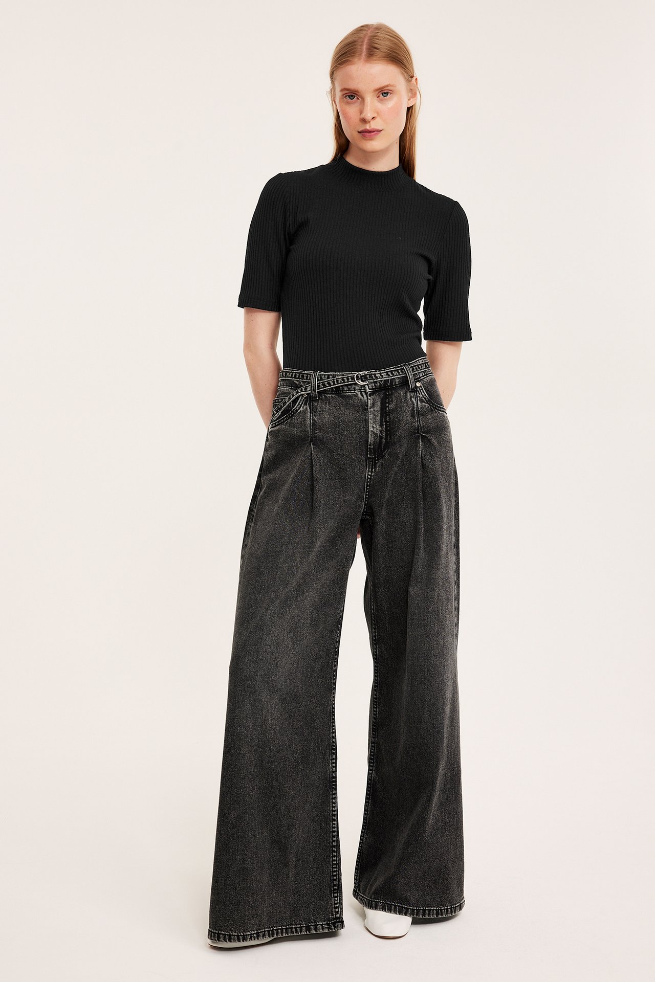 Framsida av kvinna i svart ribbad kortärmad polotopp instoppad i mörkgrå vida jeans.