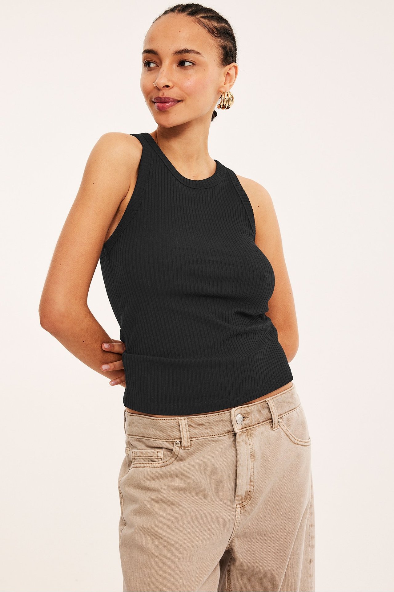 Frontvisning av en kvinne i svart ribbet singlet, beige jeans og gulløredobber.