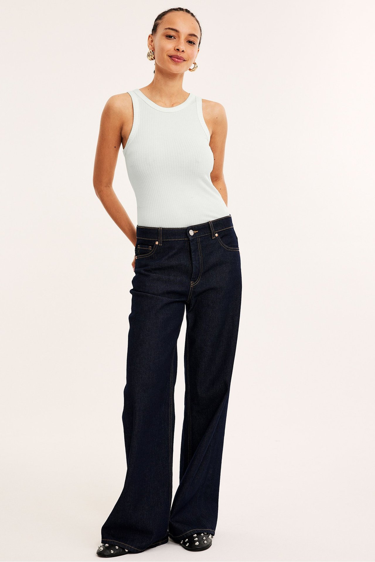 Framifrån: Kvinna i offwhite ribbat linne, mörkblå vida jeans och svarta nitprydda platta skor.