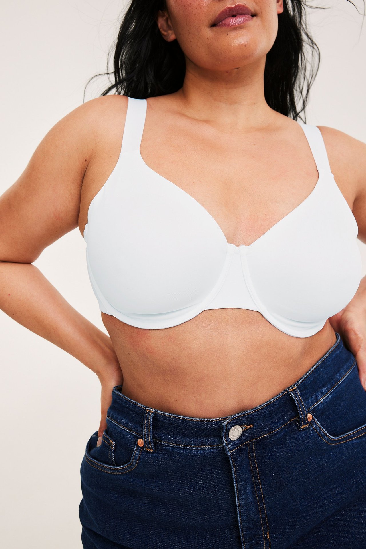 Lähikuva edestä: nainen yllään valkoiset topattomat kaarituelliset minimizer-rintaliivit ja tummansiniset farkut.
