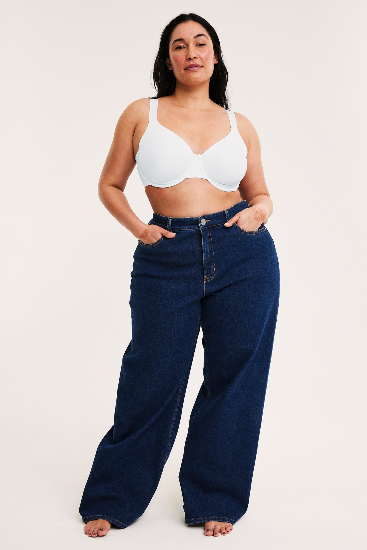 Frontvy av en kvinna i en vit bygel-minimizer-bh och mörkblå vida jeans.