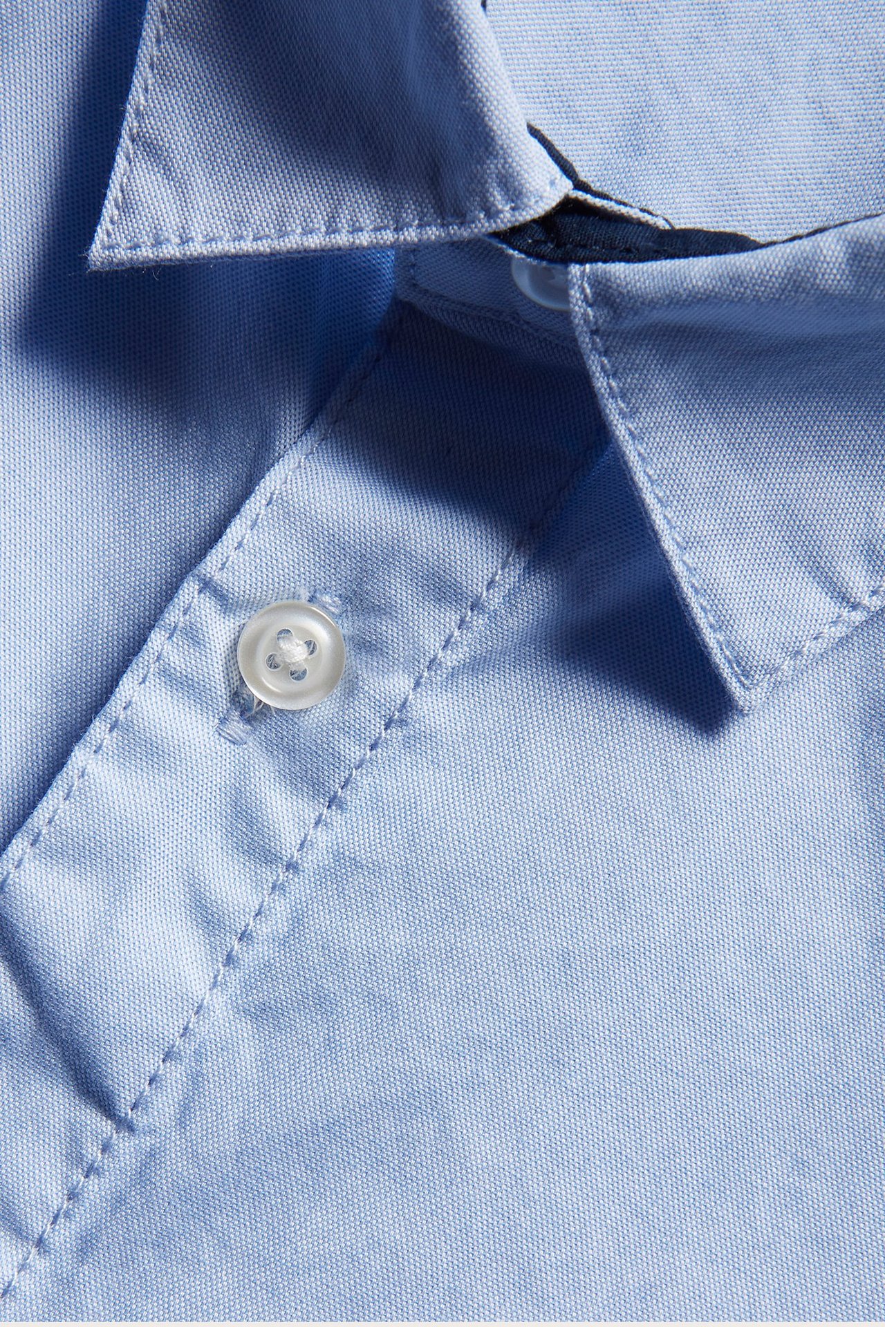Zbliżenie jasnoniebieskiej koszuli oxford z białym guzikiem na plisie i kołnierzyku.