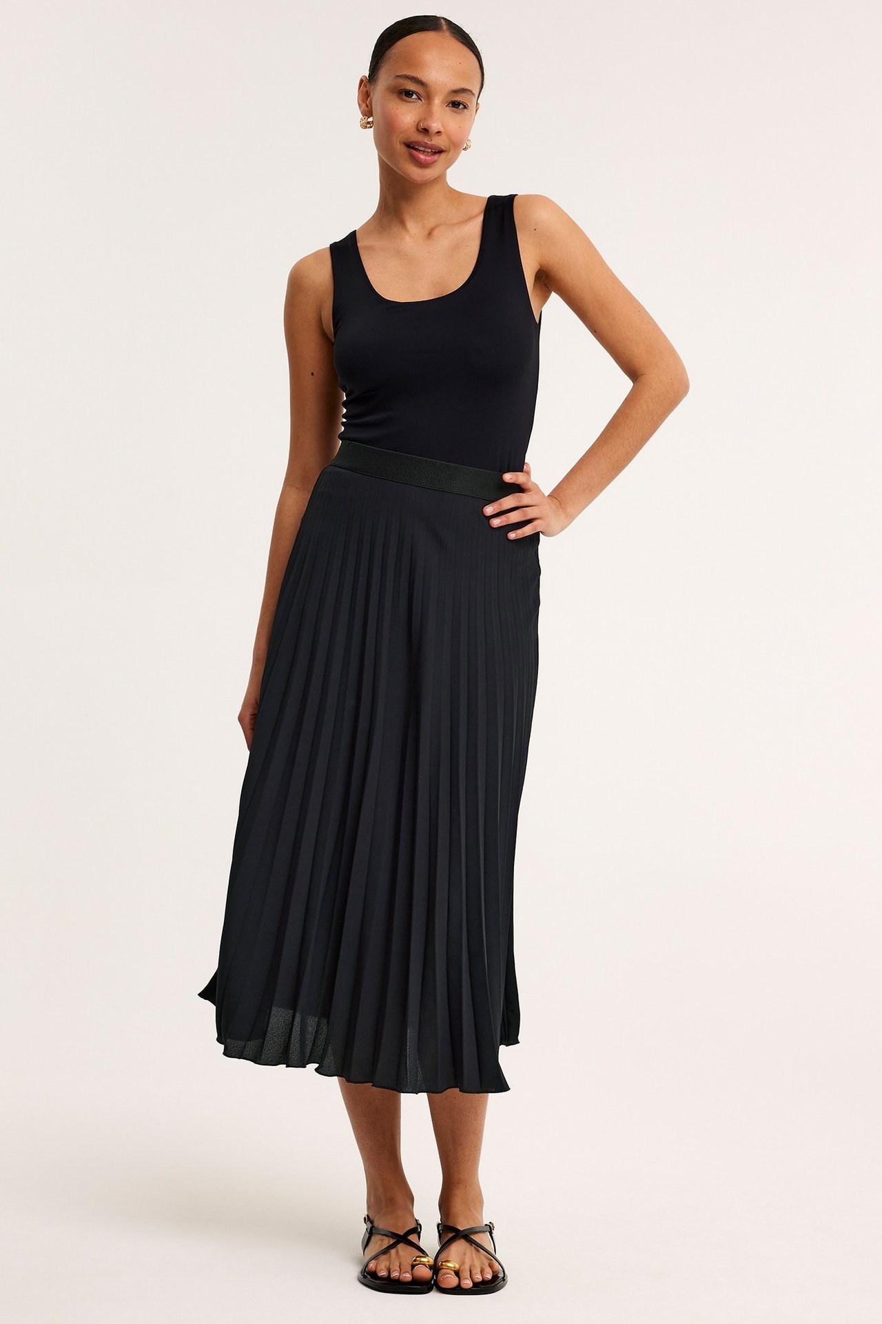 Widok z przodu: Kobieta w czarnej plisowanej spódnicy z gumką, czarnym topie, czarnych sandałach i złotej biżuterii.