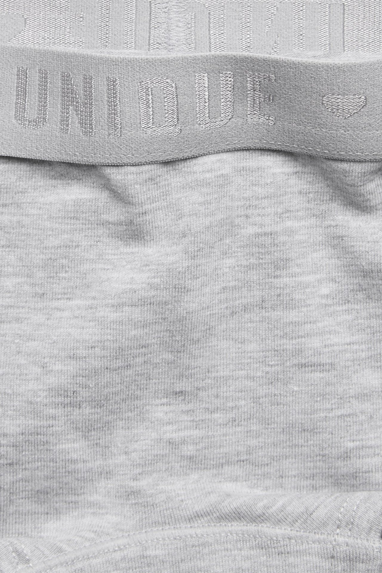 Lähikuva edestä lasten vaaleanharmaista bokserishortseista, joissa on joustava vyötärönauha 'UNIQUE'-tekstillä ja sydämellä.