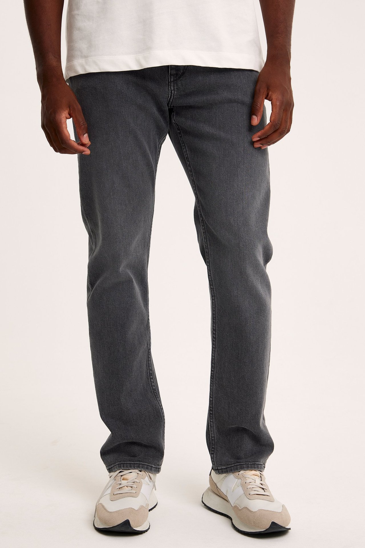 Framifrån, en man bär gråa regular fit denimjeans, en vit t-shirt och beige-vita sneakers.