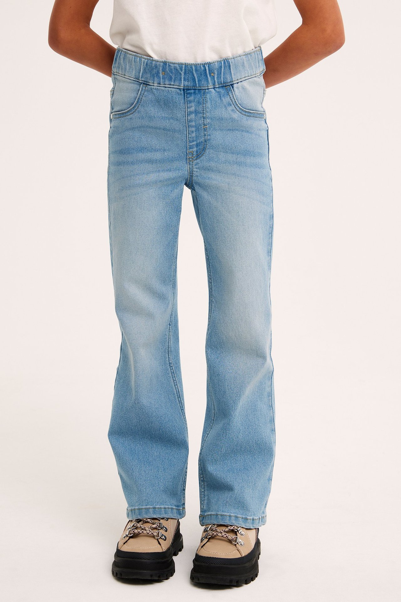 Edestäpäin, lapsi yllään vaaleansiniset bootcut-denimleggingsit, valkoinen t-paita ja beiget paksupohjaiset kengät.