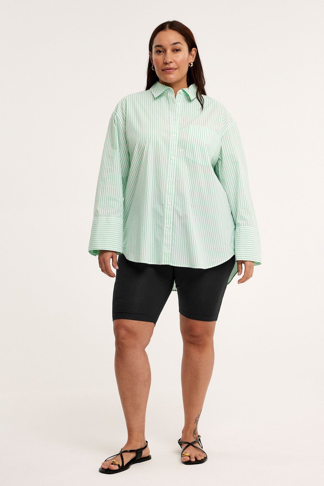 Edestä: nainen mustissa jersey-pyöräilyshortseissa, raidallisessa ylisuuressa paidassa ja mustissa sandaaleissa.