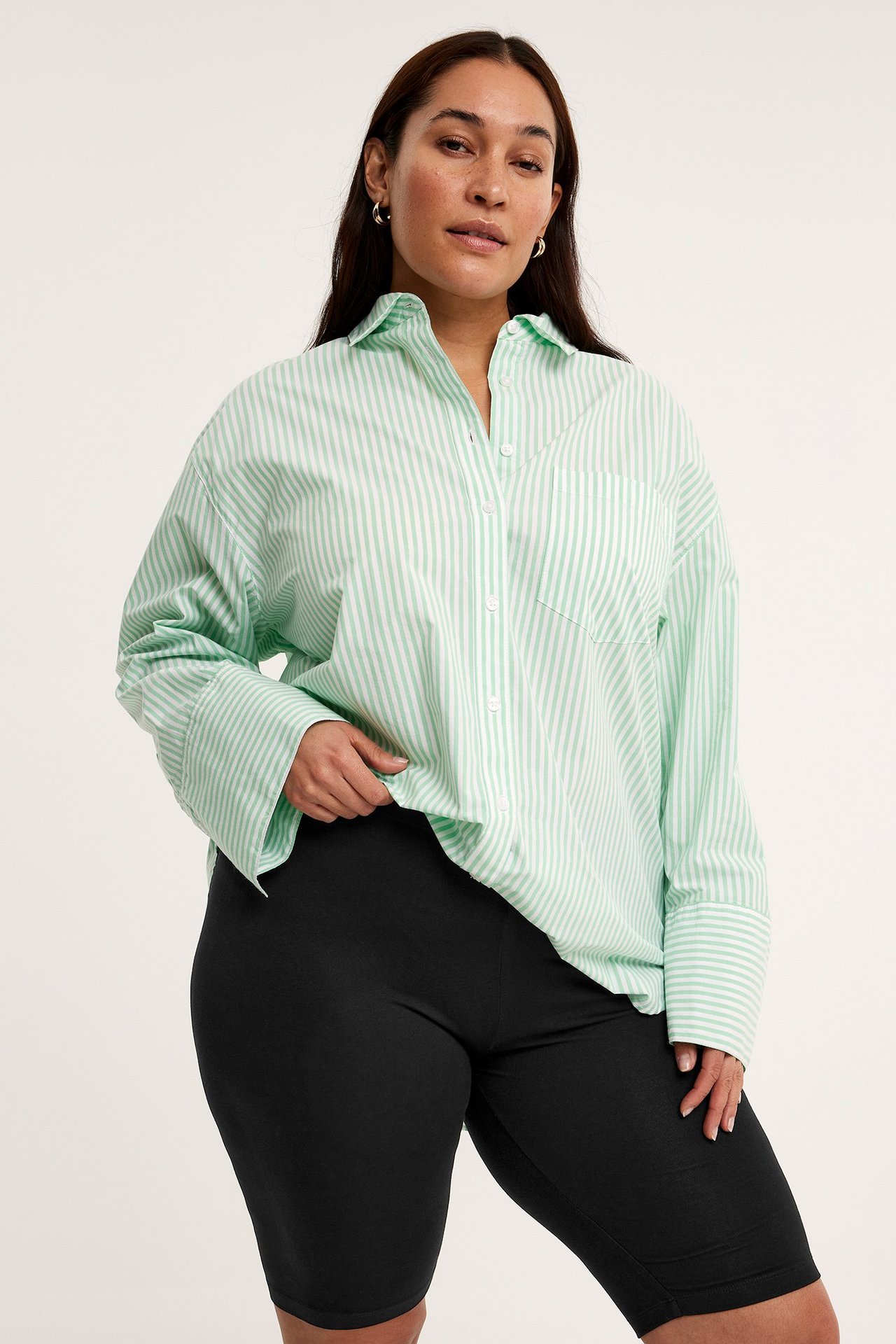 Widok z przodu kobiety w czarnych dżersejowych szortach rowerowych i oversize'owej jasnozielonej koszuli w paski.