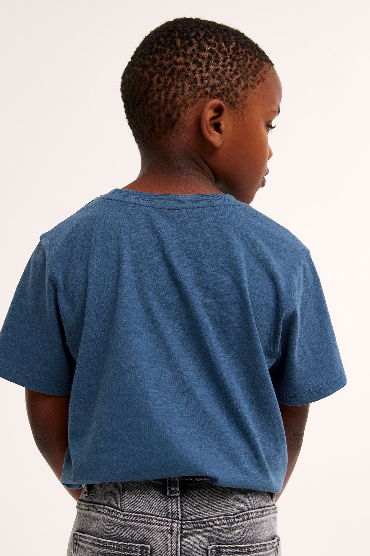 Bakifrån, ett barn i en mörkblå bomulls-t-shirt och grå jeans.