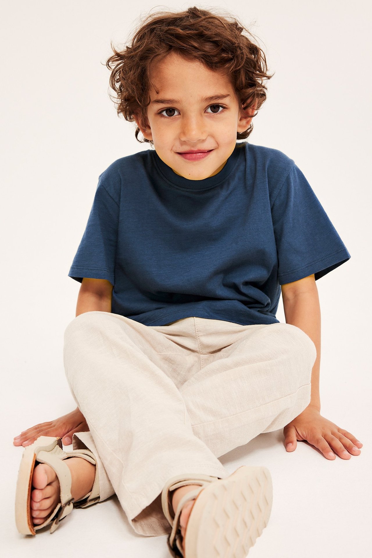 Edestä kuvattu lapsi yllään tummansininen puuvilla-T-paita, vaaleanbeiget housut ja beiget sandaalit.