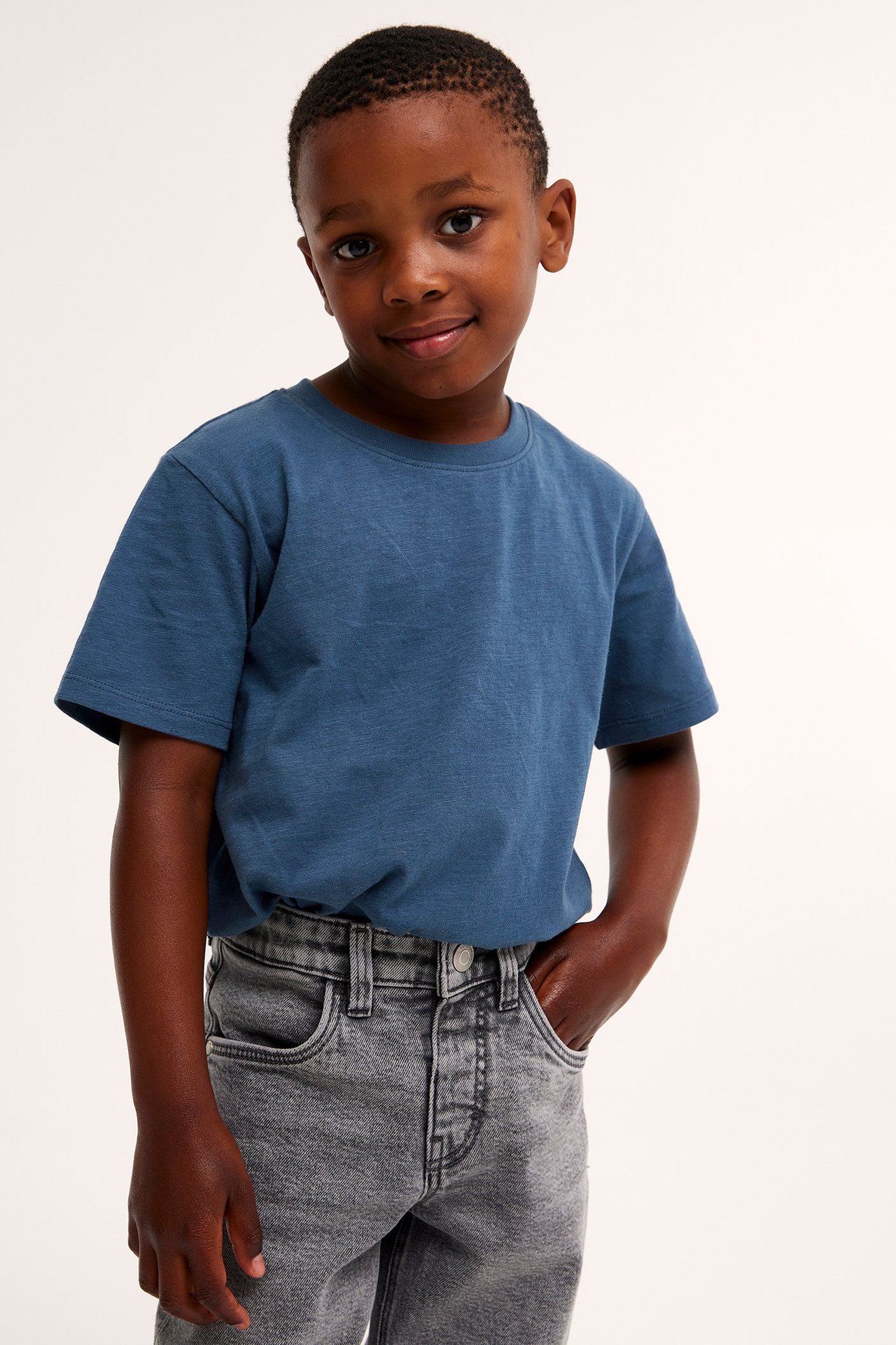 Edestä: lapsi tummansinisessä lyhythihaisessa puuvillat-paidassa ja harmaissa farkuissa.