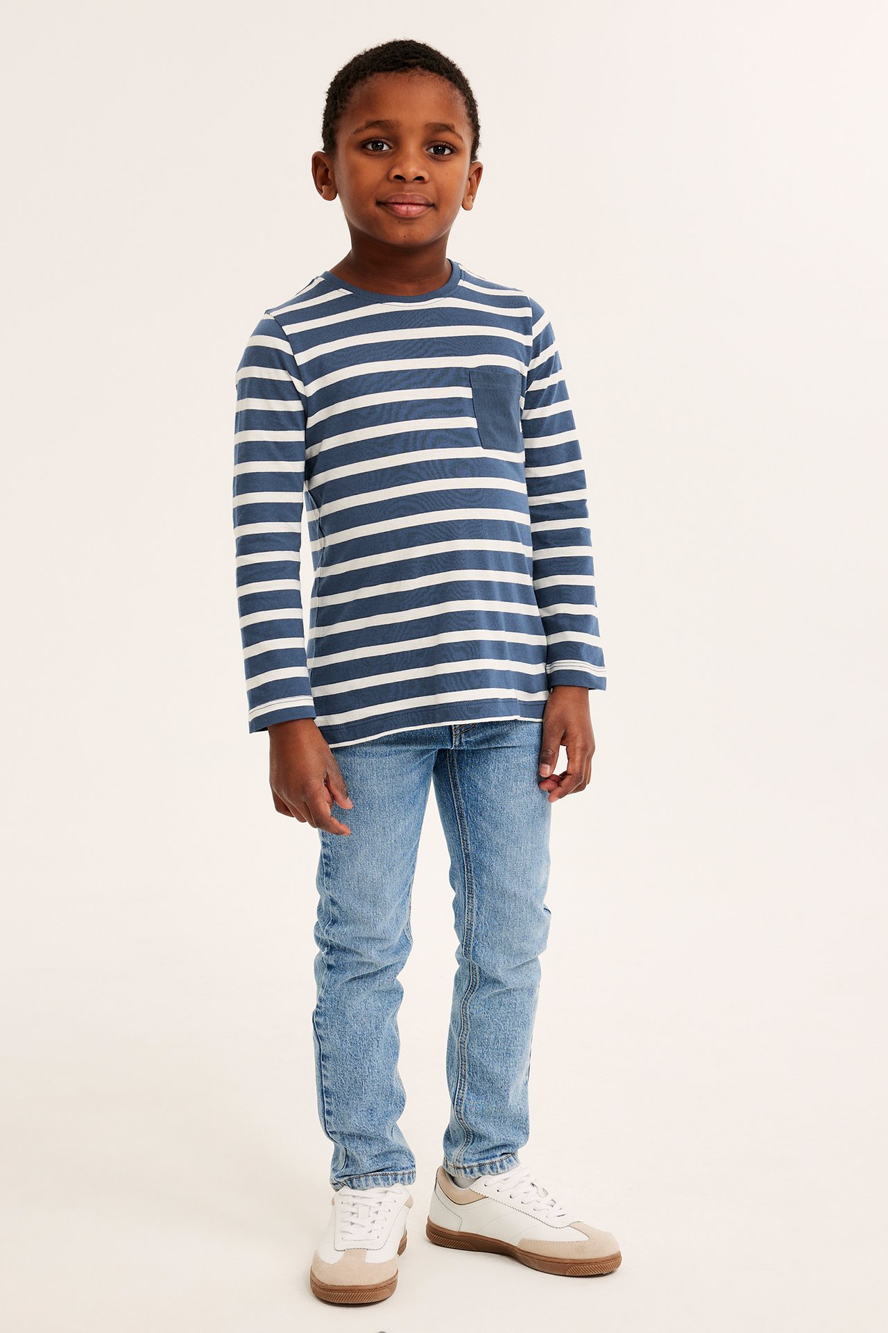 Framifrån, ett barn i mörkblå- och vitrandig långärmad t-shirt, ljusblå jeans och vita sneakers.