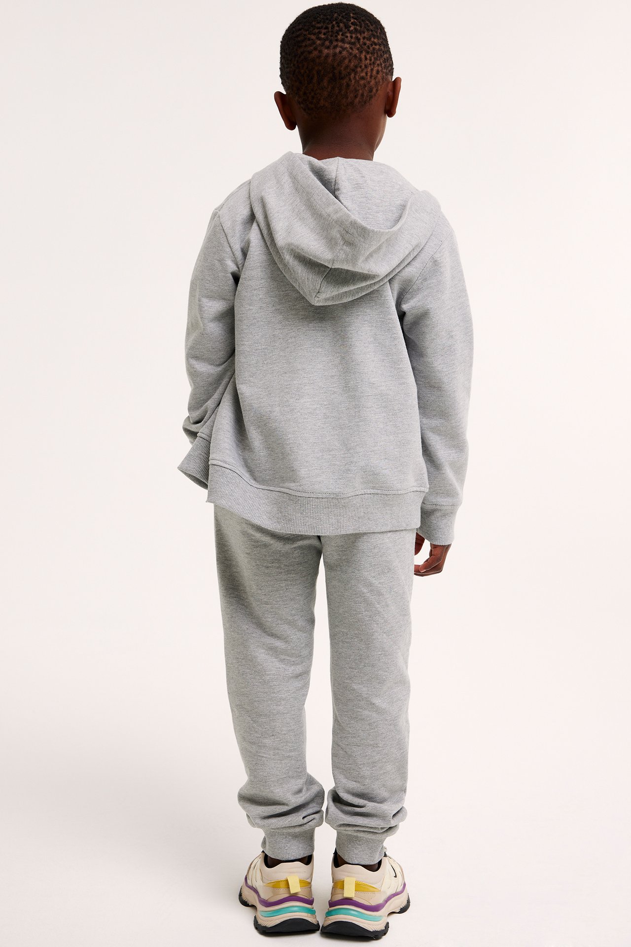 Bakifrån, ett barn i melerade grå mjukisbyxor med ribbade muddar, matchande huvtröja och sneakers.