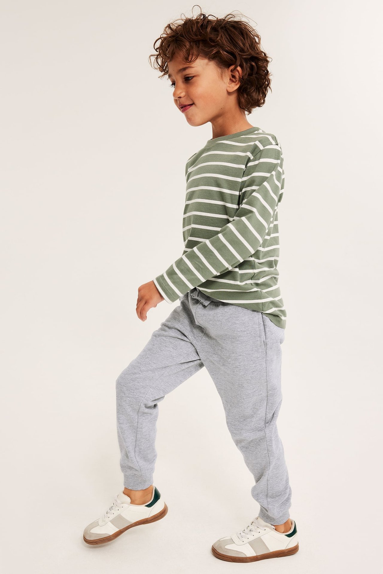 Sidovy av ett barn i melerade grå mjukisbyxor, en randig långärmad tröja och vita sneakers.