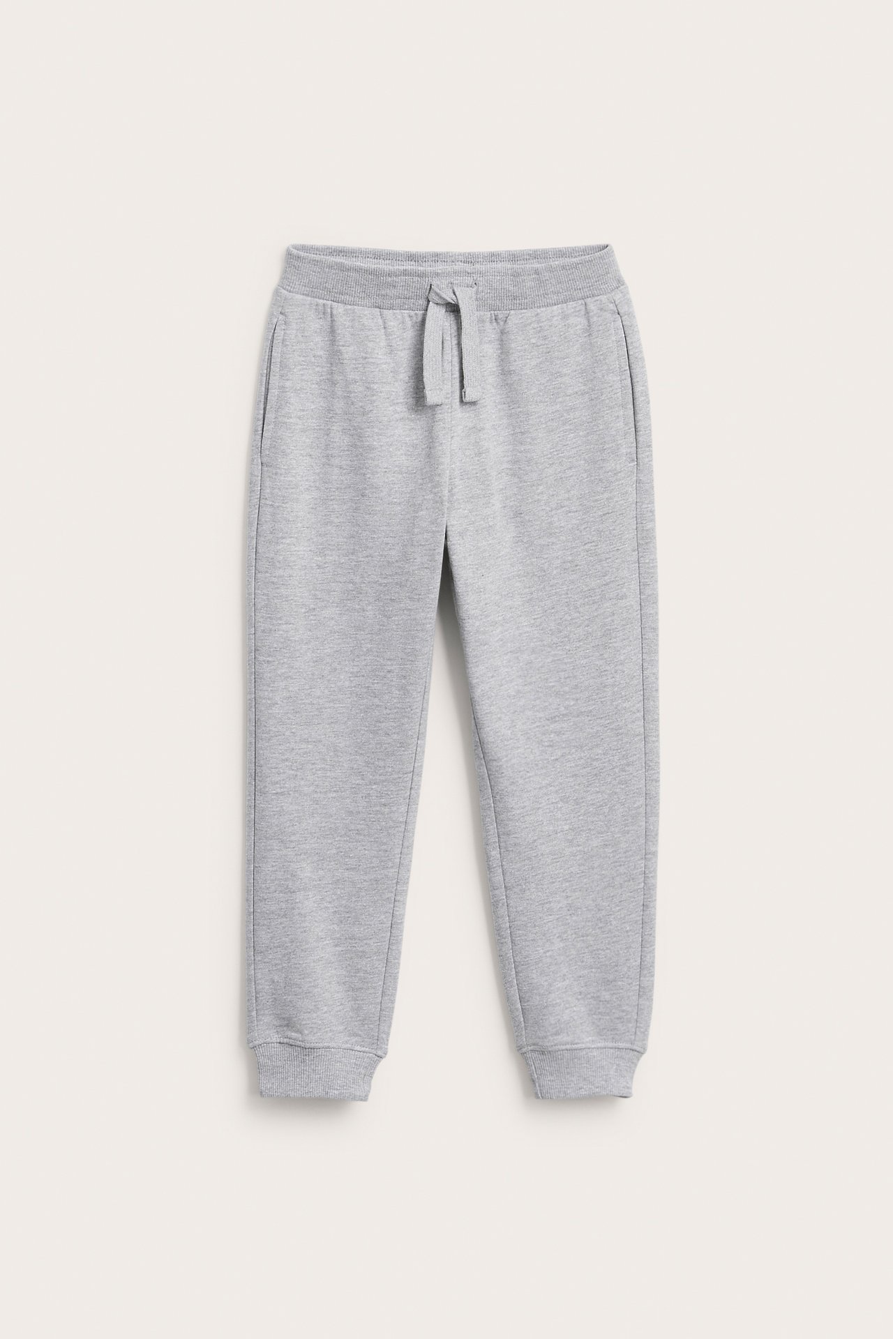 Lasten harmaat melange-collegehousut edestä: joustava nyörivyötärö, resorit lahkeissa ja sivutaskut.