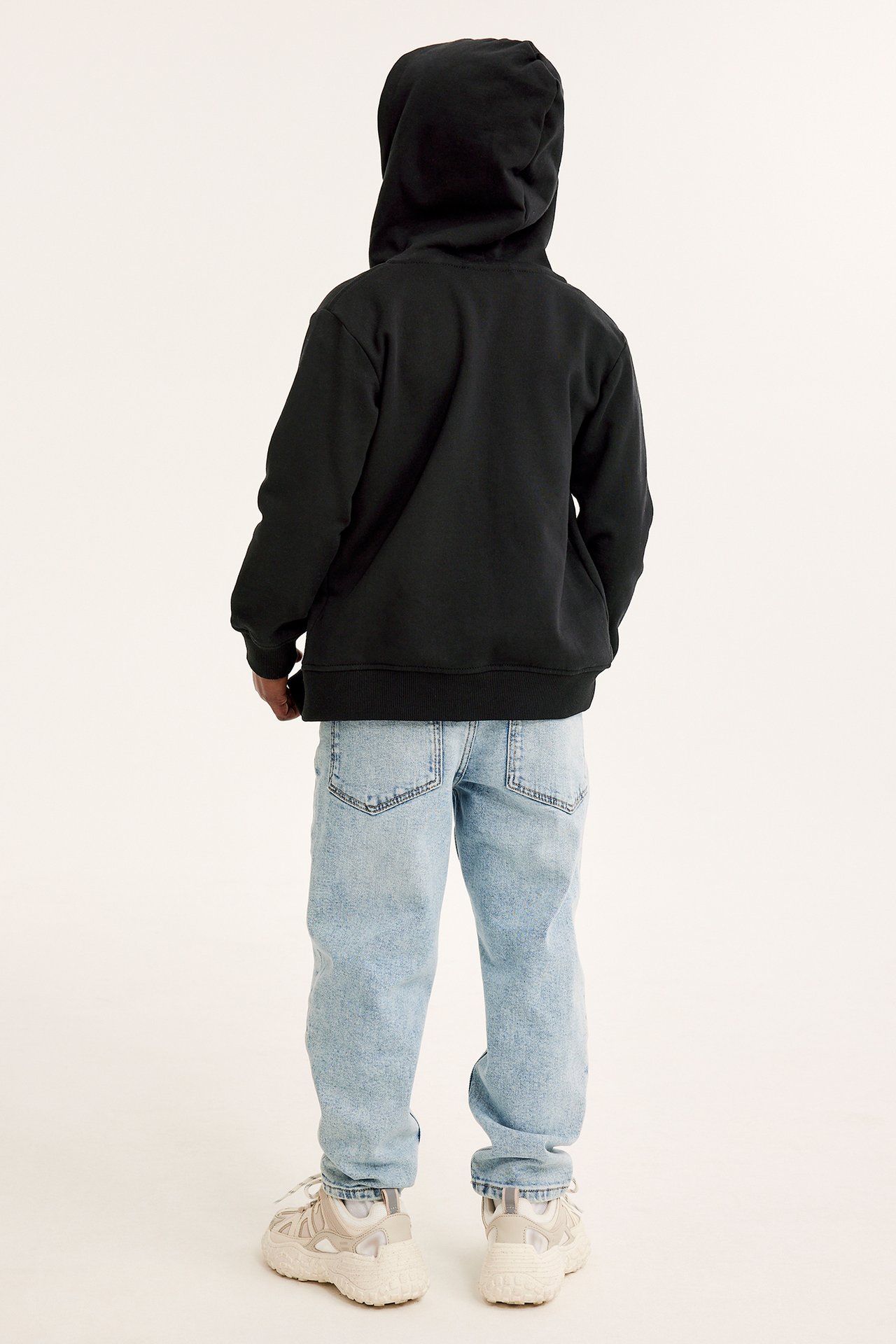 Bakifrån: ett barn i svart bomullshoodie med dragkedja, ljusblå jeans och ljusbeige sneakers.