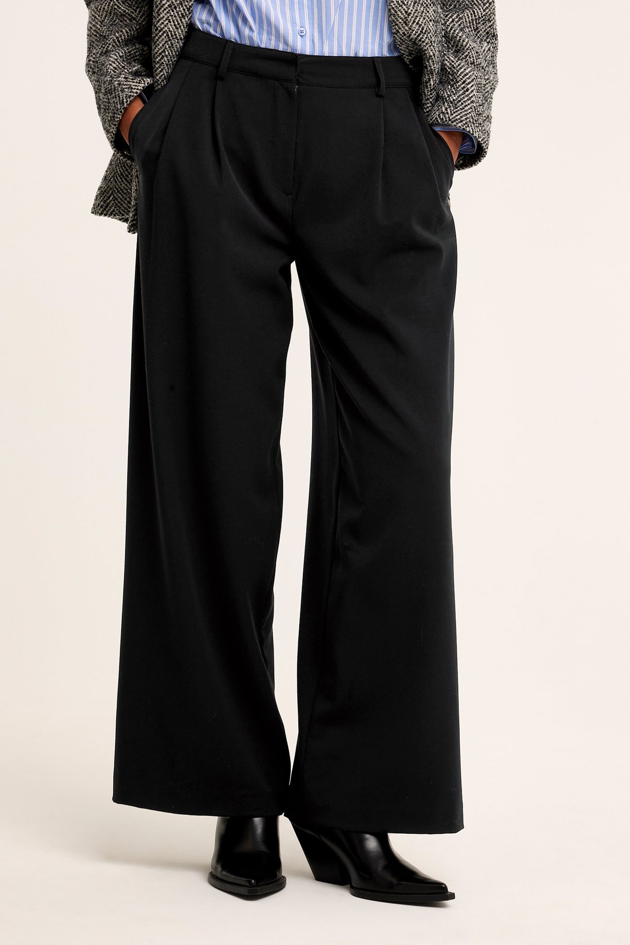 Kvinne i svarte vide plisserte bukser, stripete skjorte, blazer og svarte støvler.