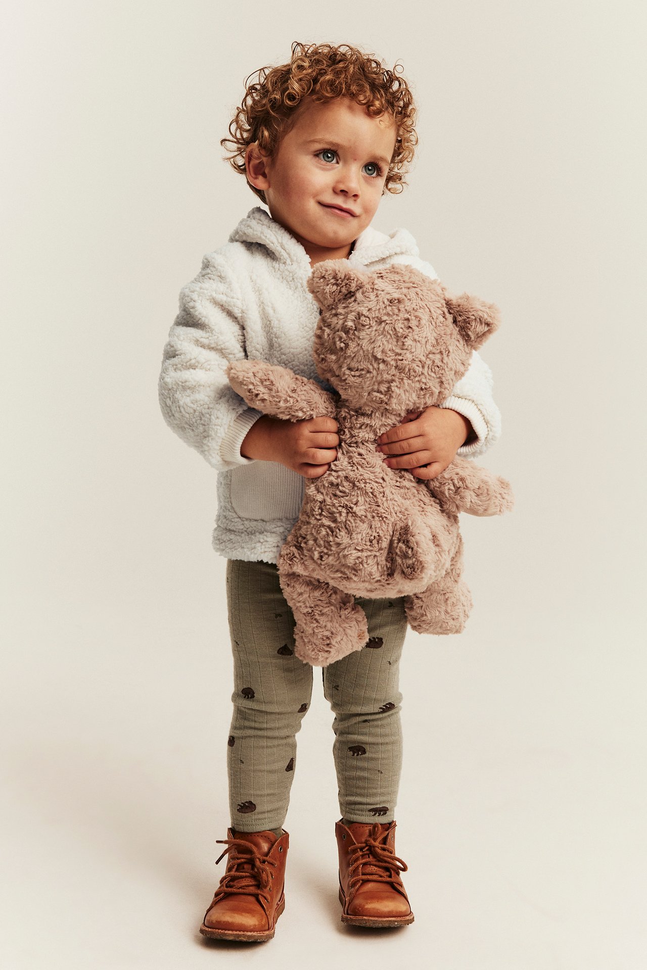 Forfra: Barn i hvit lodden hettegenser, olivenfargede leggings, brune sko, holder en brun bamse.