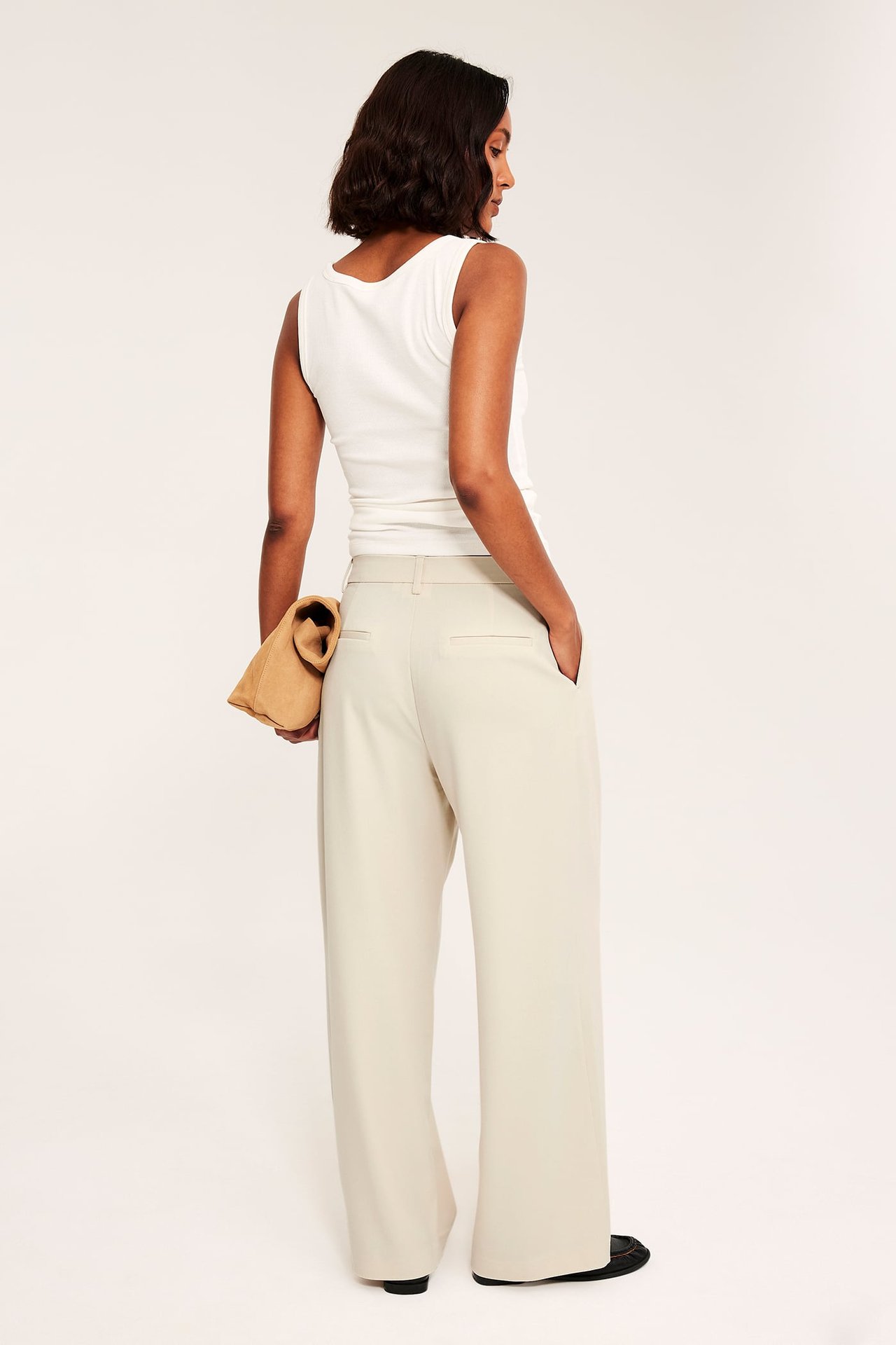 Bakifrån kvinna i ljusbeige vida byxor, vit ärmlös topp och brun clutch.
