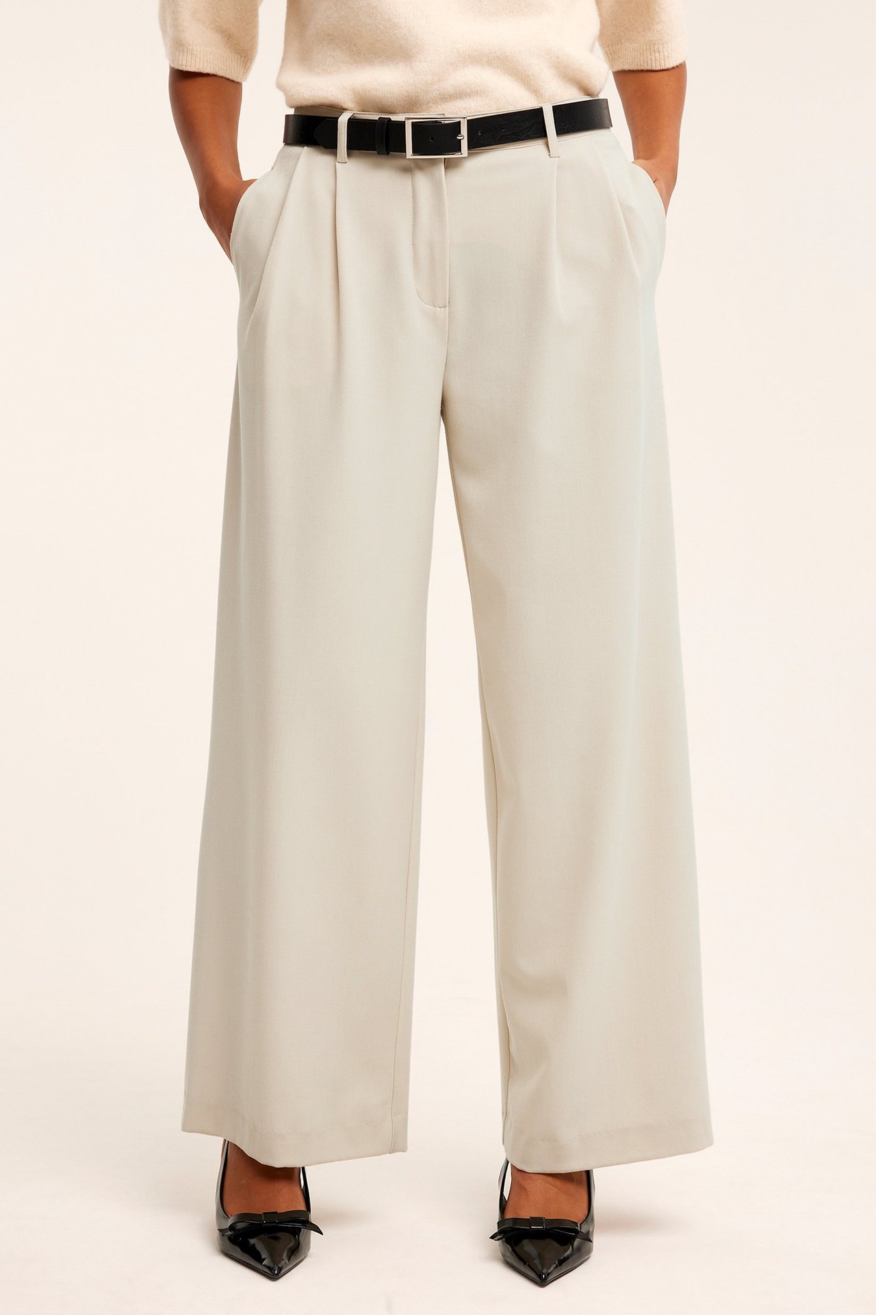 Frontbilde: kvinne i lys beige vide dressbukser med legg, svart belte, lys beige genser og svarte hæler.