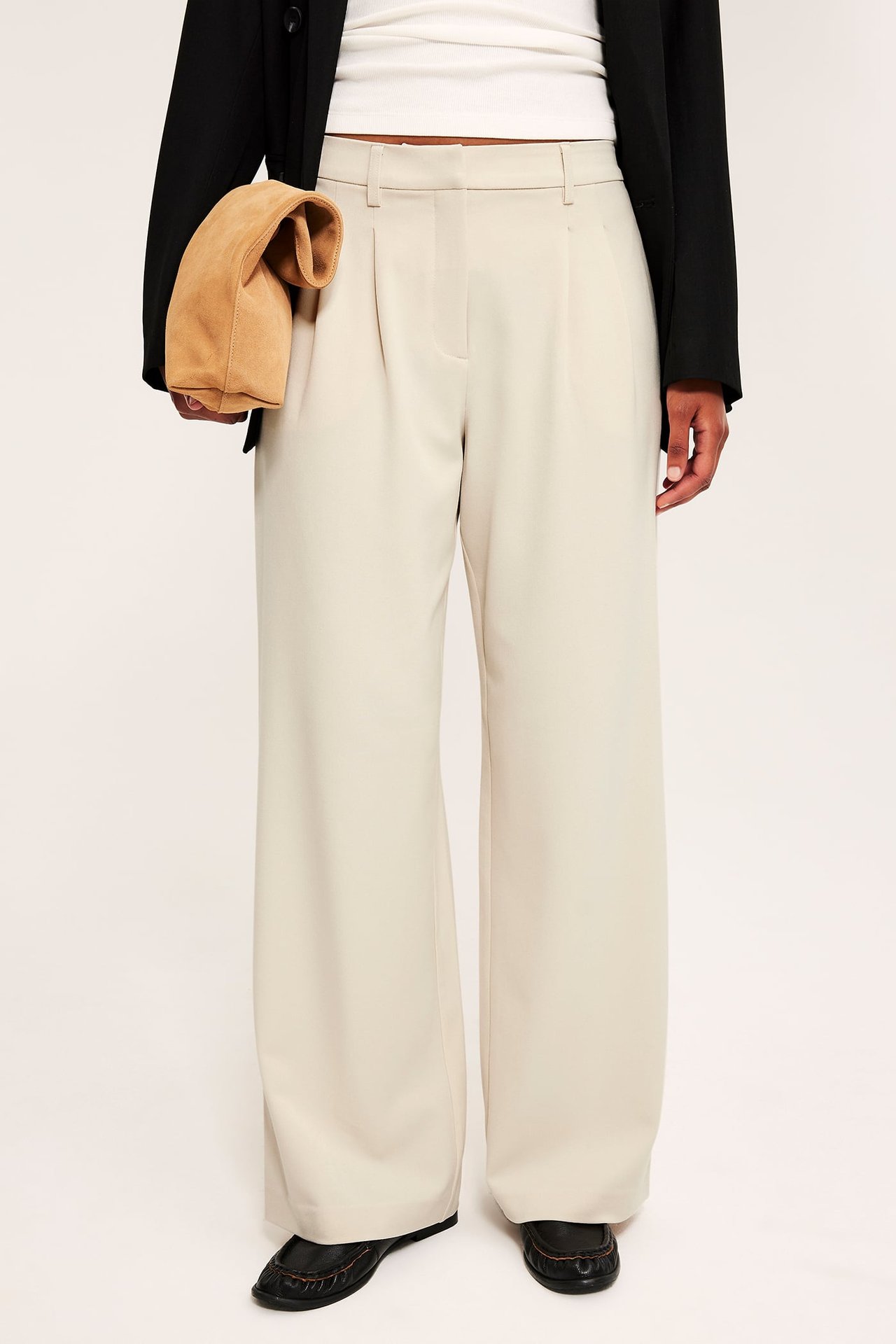 Dame i lysebeige vide bukser, svart blazer, beige semsket clutch og svarte loafers.