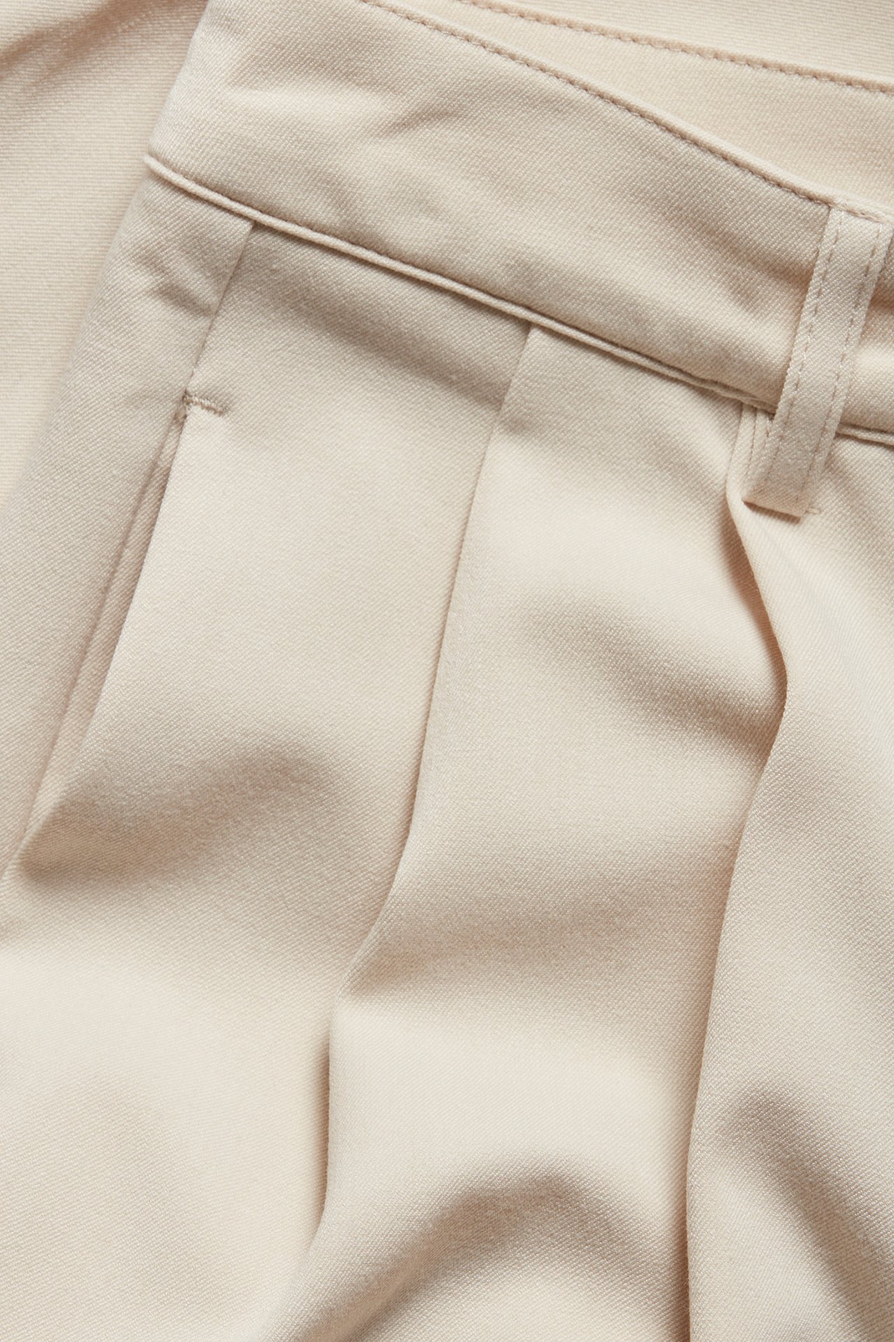 Zbliżenie beżowych spodni garniturowych z zakładkami, przednimi kieszeniami i szlufką.