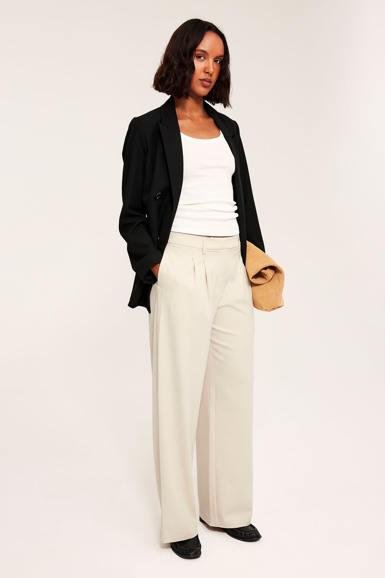 Frontbilde: kvinne i lys beige vide bukser, sort blazer, hvit topp, holder brun veske.
