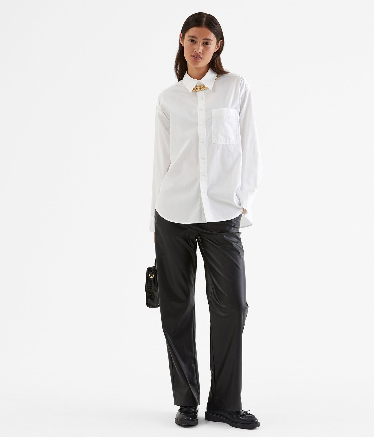 Kobieta w białej koszuli z bawełny ze złotym łańcuchem, czarnych spodniach i mokasynach, trzymająca czarną torebkę.