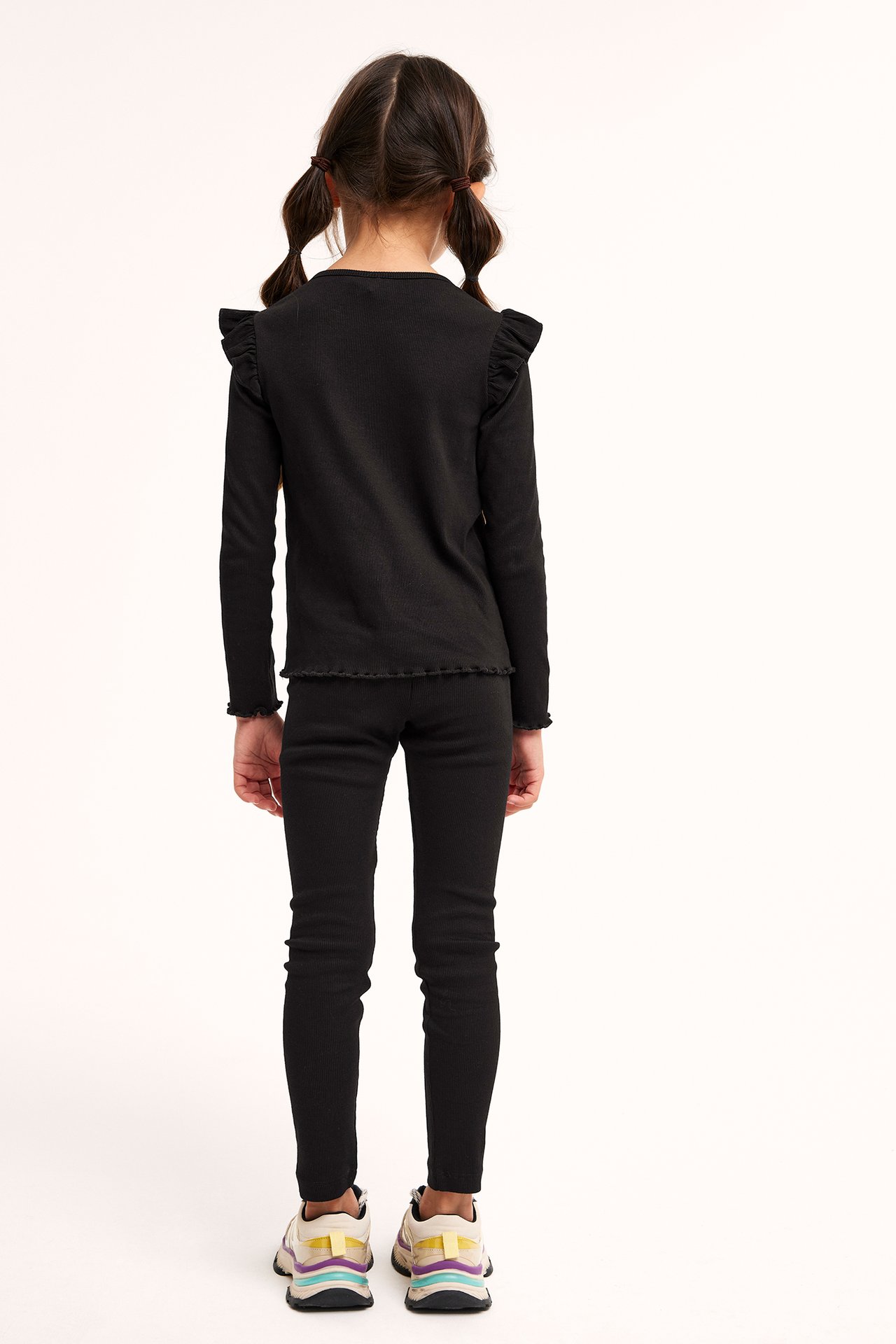 Bakifrån på ett barn i svarta ribbade leggings, svart volangtopp och färgglada sneakers.