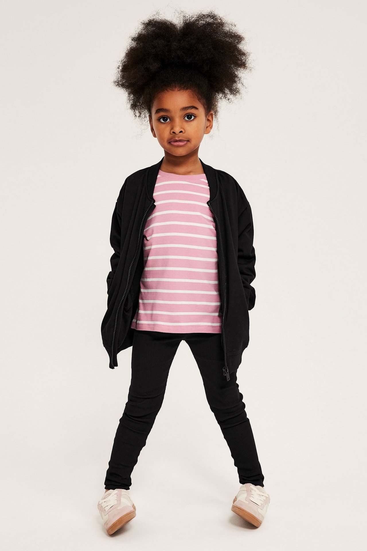 Z przodu: dziecko w czarnych prążkowanych legginsach, różowej bluzce w paski, otwartej czarnej kurtce bomberce i trampkach.