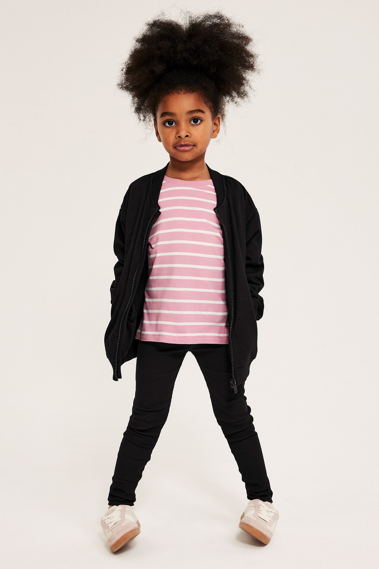Framifrån, ett barn i svarta ribbade leggings, en rosa randig t-shirt och en öppen svart bomberjacka.