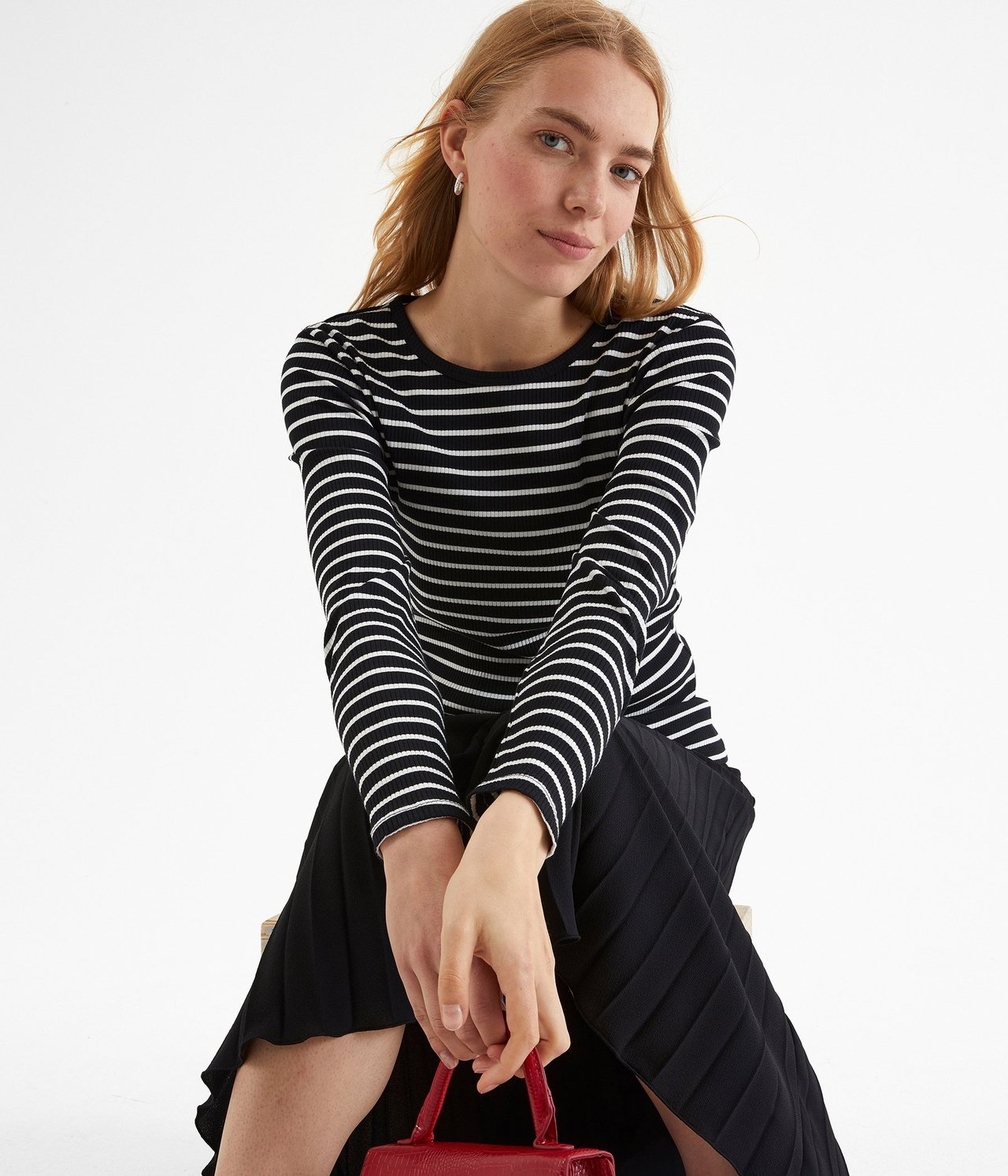Forfra: kvinne i svart- og hvitstripet ribbet langermet topp, svart plissert skjørt, holder rød håndveske.