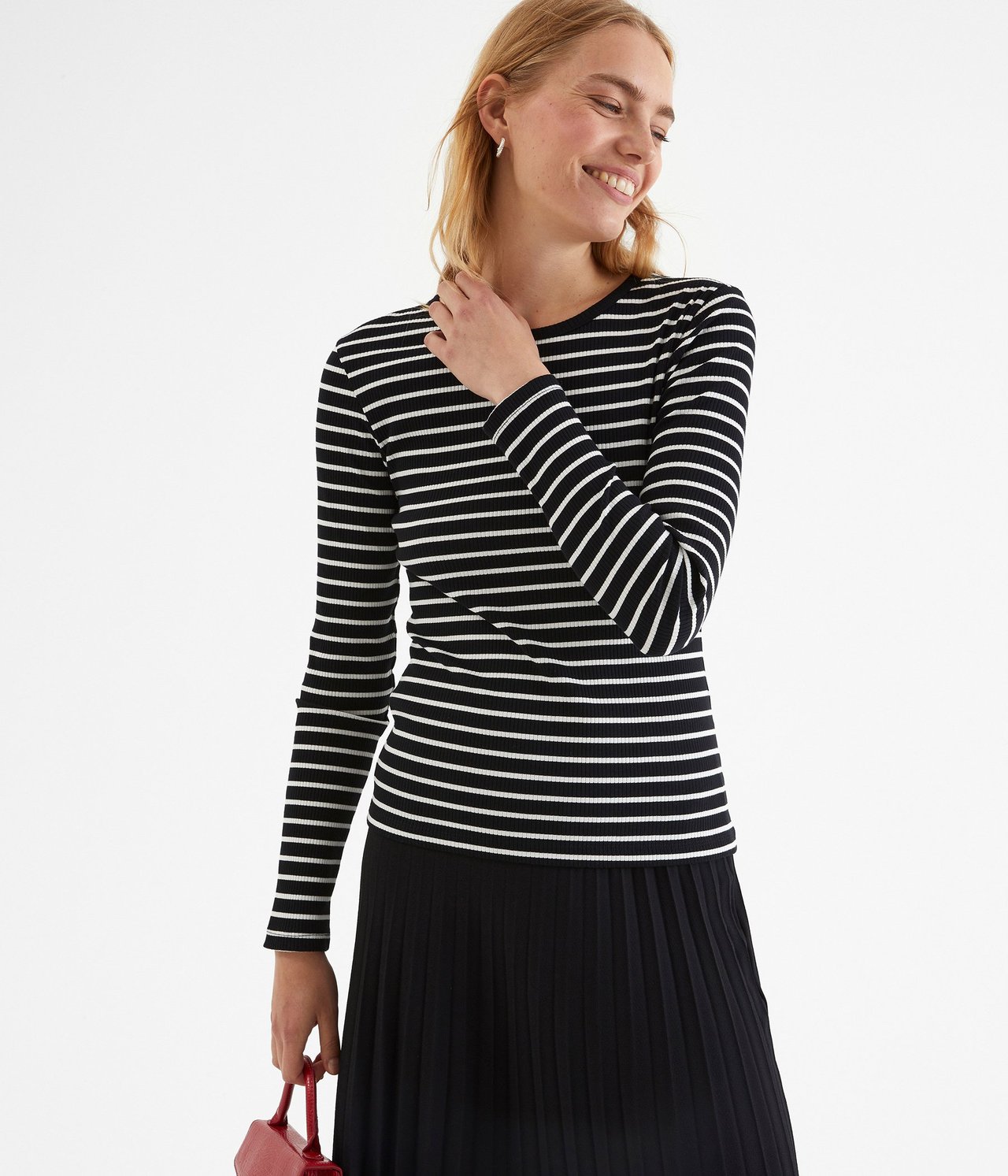 Forfra: Kvinne i svart- og hvitstripet ribbet langermet topp, svart plissert skjørt, holder en rød veske.