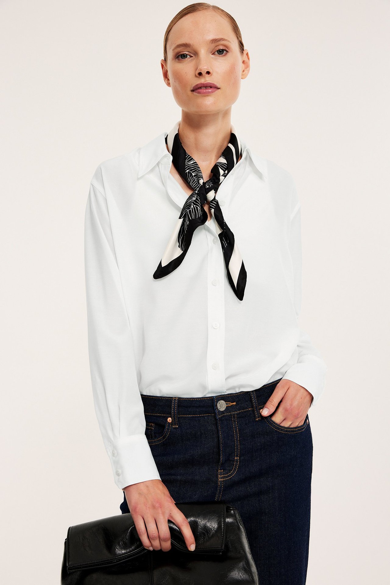 Naisen etukuva: valkoinen oversized-paita, mustavalkoinen huivi, tummat farkut, musta käsilaukku.