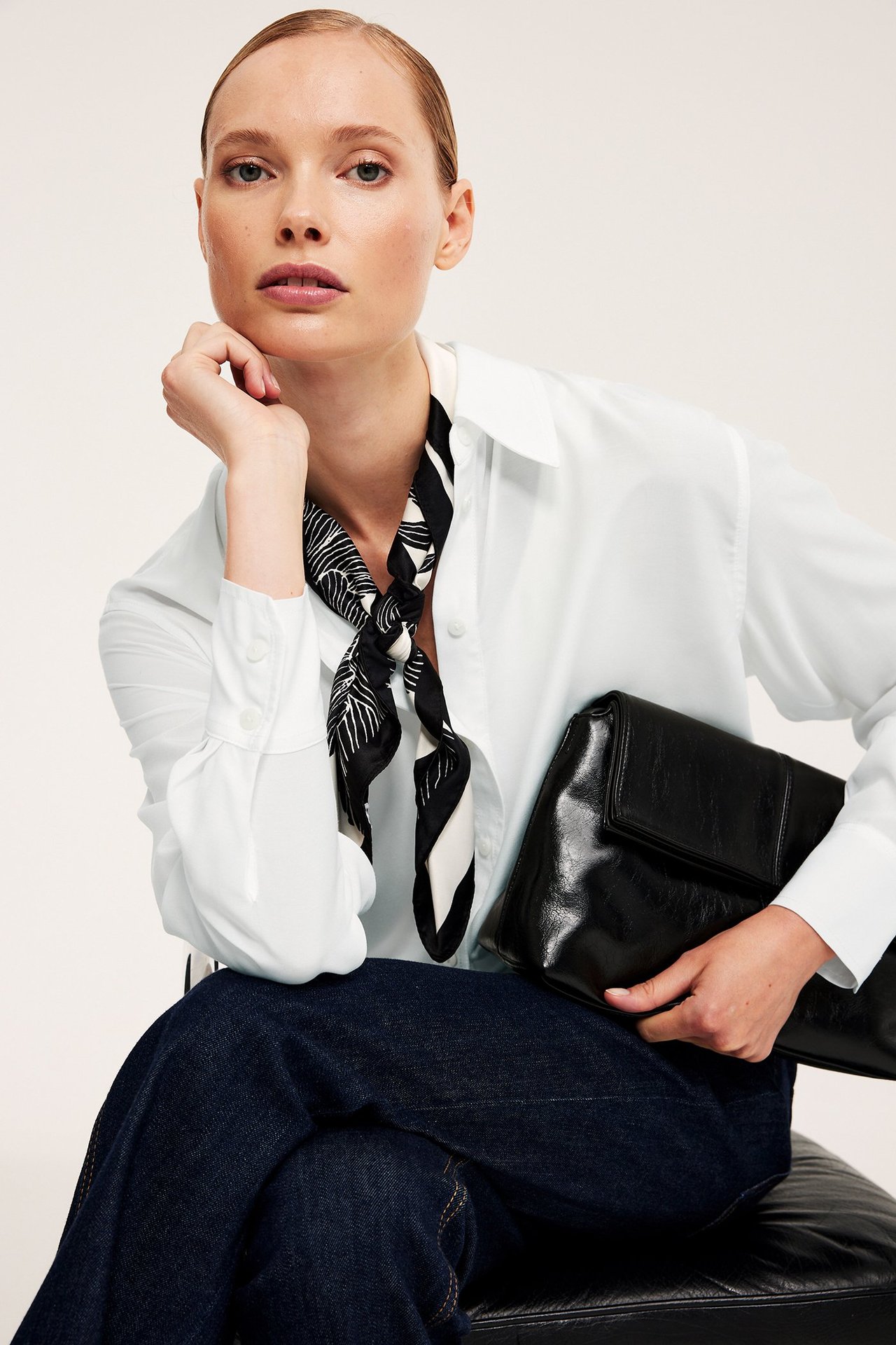 Edestäpäin nainen valkoisessa oversize-paidassa, mustavalkoisessa huivissa, tummansinisissä farkuissa, pitäen mustaa kytkinlaukua.